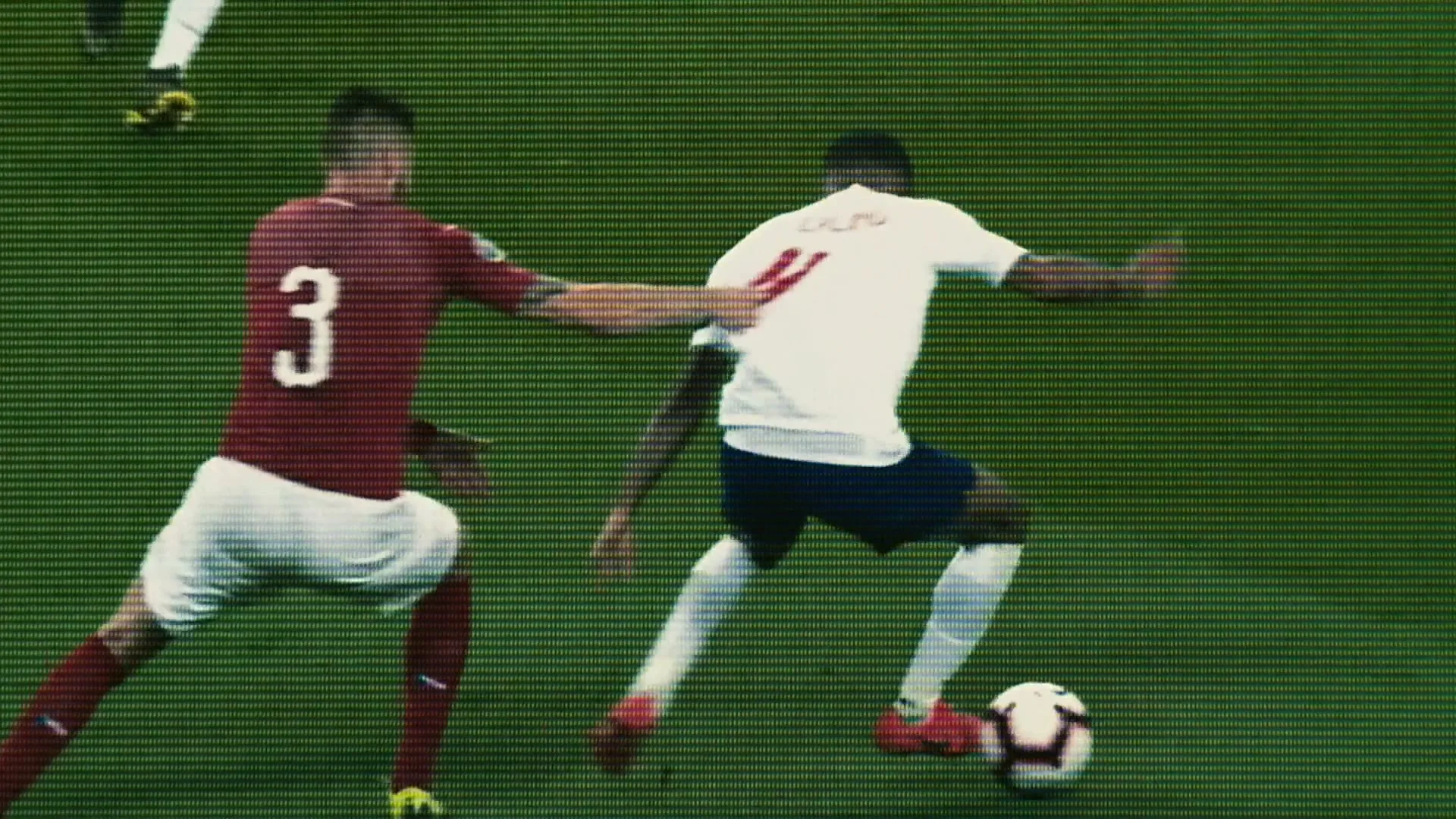 Two male soccer players, one in a white jersey and one in a red jersey, compete for the ball on a green field.