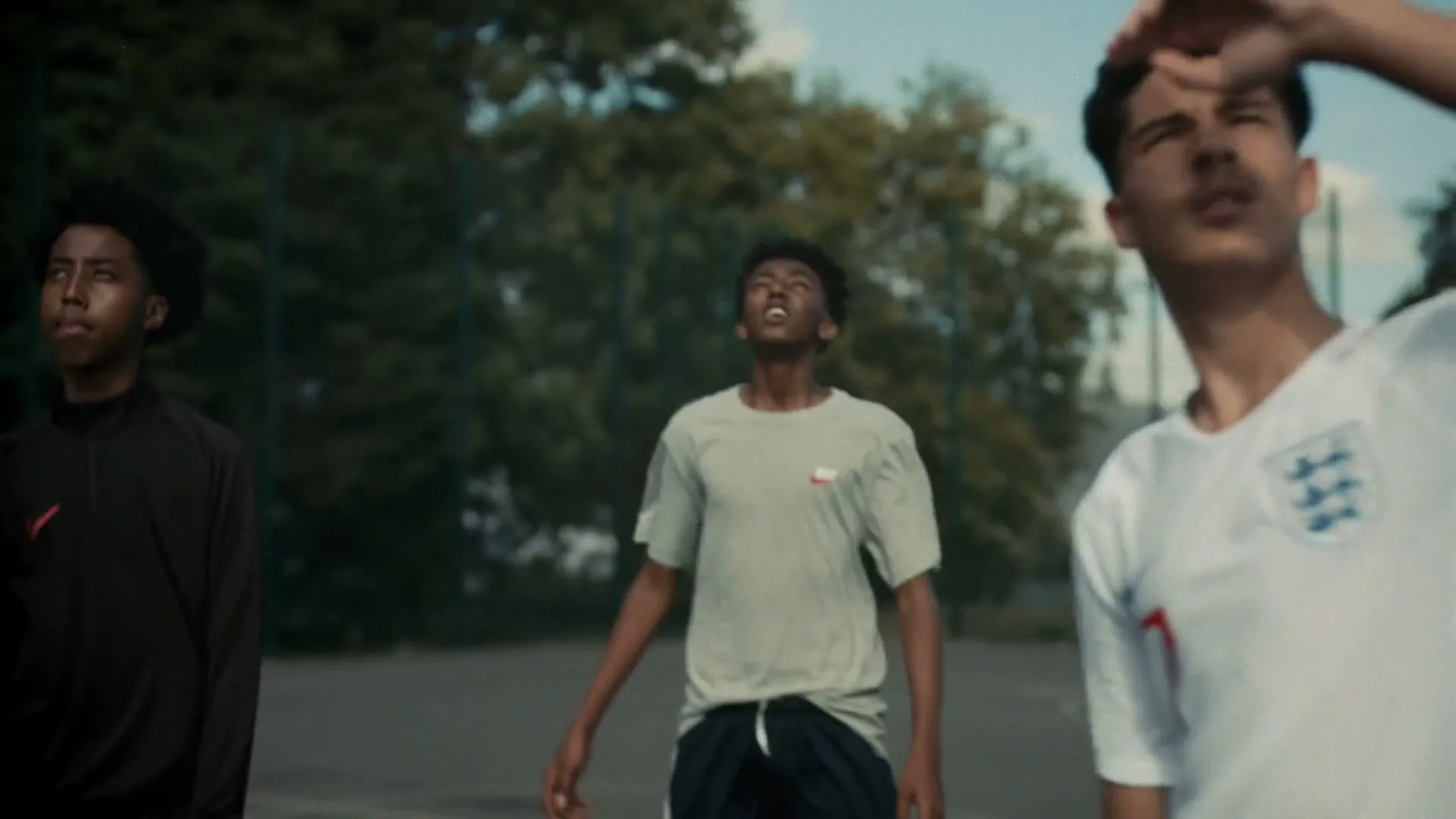 Three young male soccer players stand on an outdoor court, looking up at something off-screen, with trees in the background.