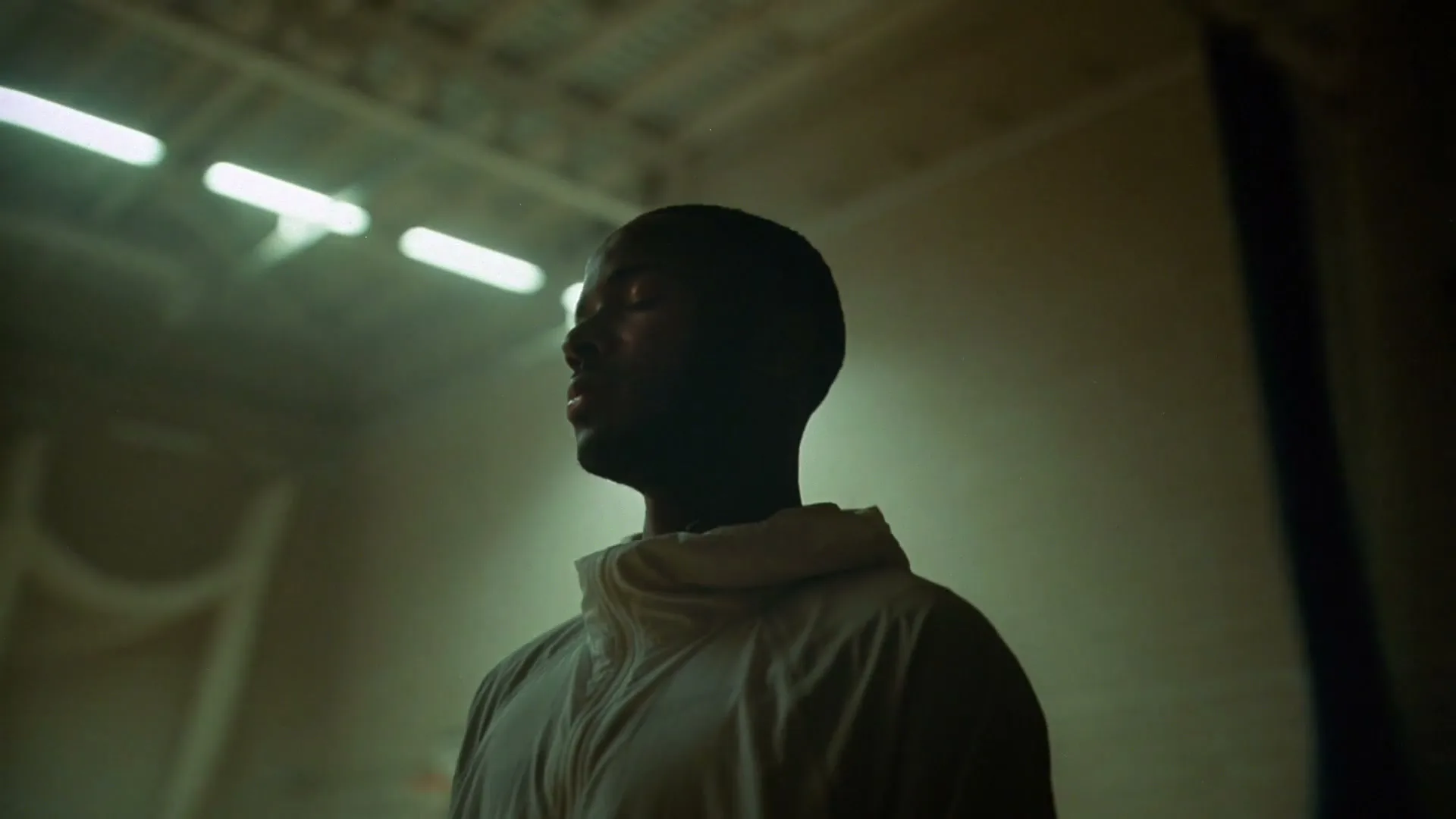 A young Black man in a light hoodie stands with his eyes closed, head slightly tilted, in a dimly lit gym.