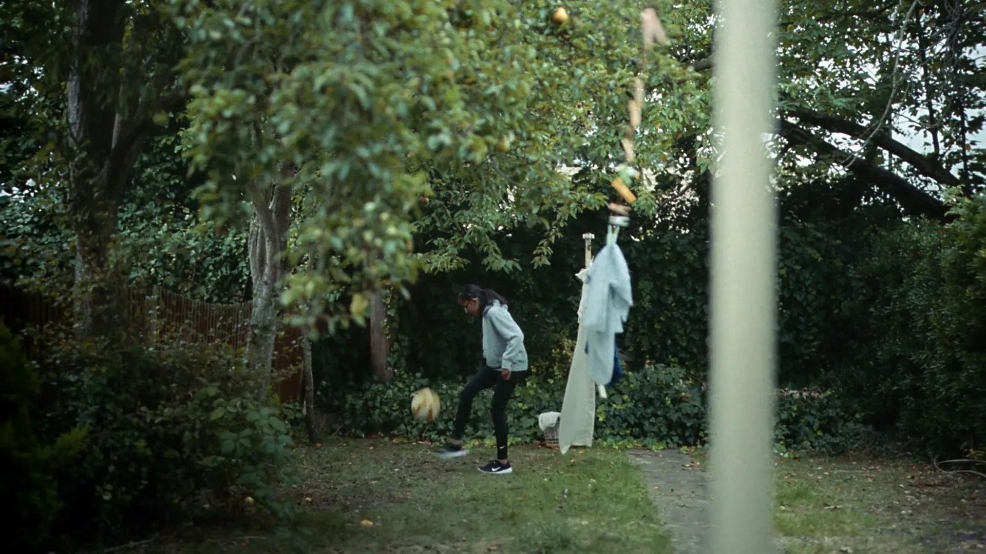 A young girl in a grey hoodie and black pants practices juggling a soccer ball in a lush green backyard during the day.