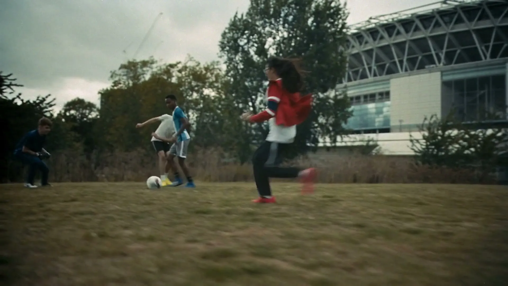 Three young people play soccer on a grassy field with a stadium in the background under an overcast sky.