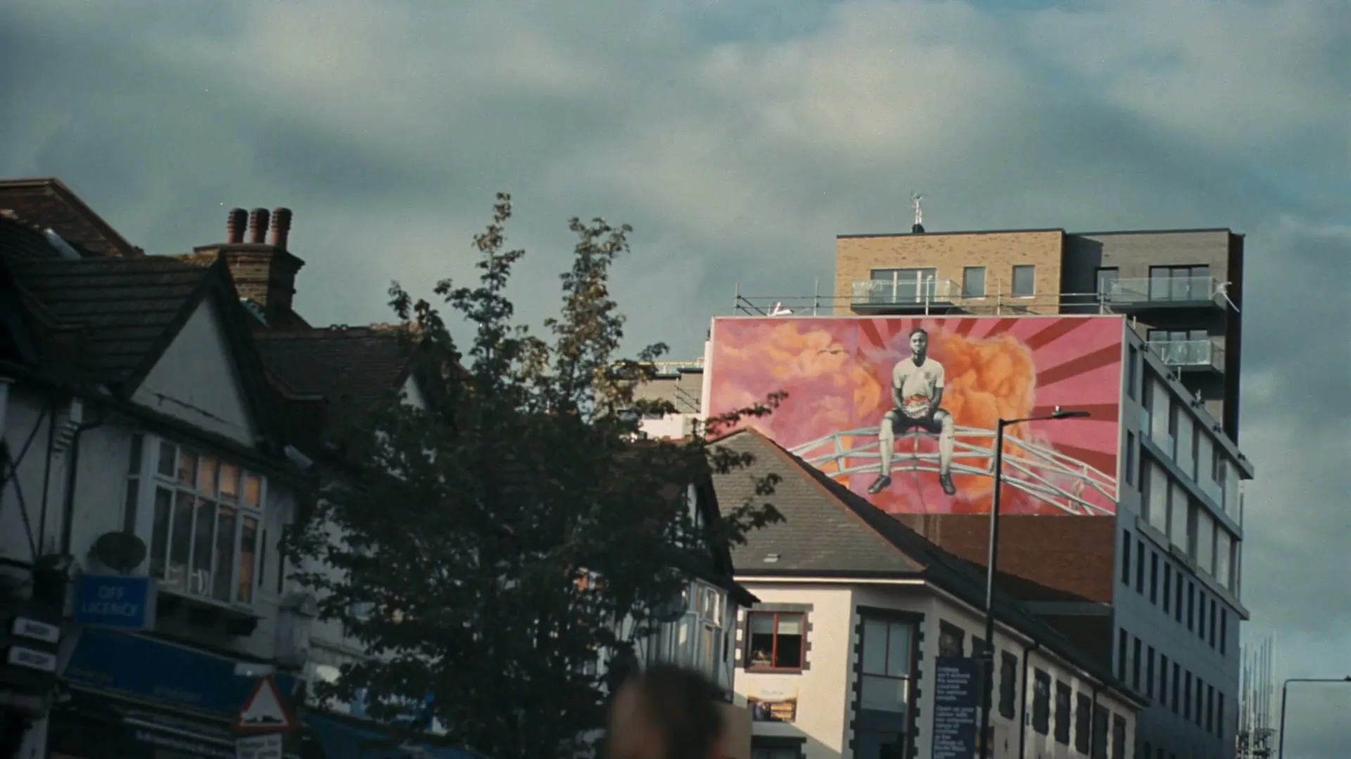 A large mural of a young Black man in a white shirt sitting on a bridge is painted on the side of a building in an urban area.