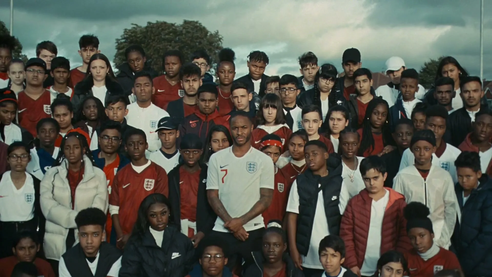 Raheem Sterling, a male soccer player, stands in the middle of a large group of diverse children and teens, all wearing soccer jerseys, on an outdoor field.