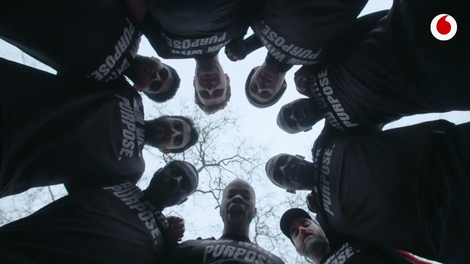 A diverse group of men in black t-shirts huddle together, looking down at the camera with determined expressions, outdoors under a cloudy sky.