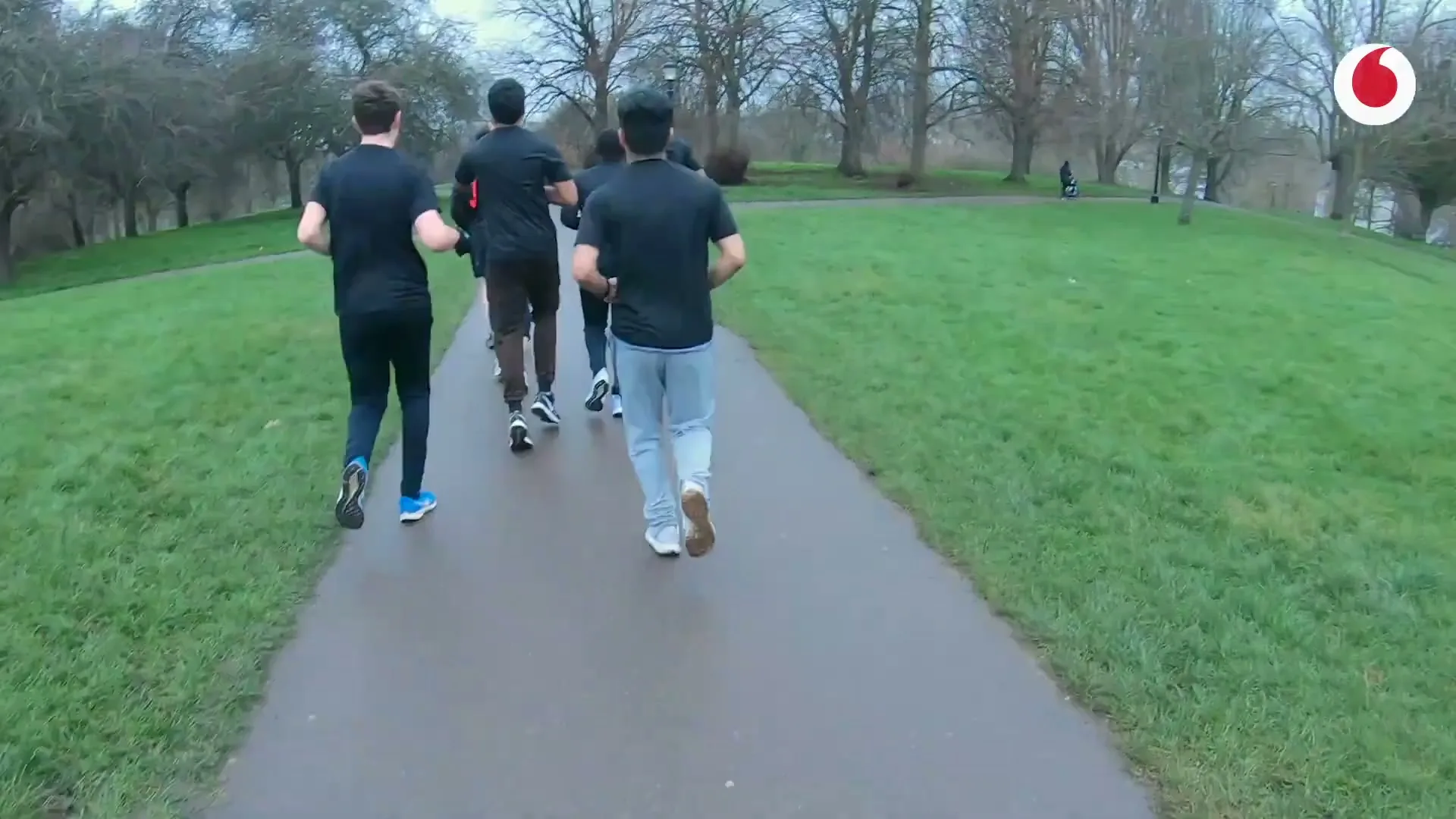Four young men in dark athletic wear run along a paved path through a park on an overcast day.