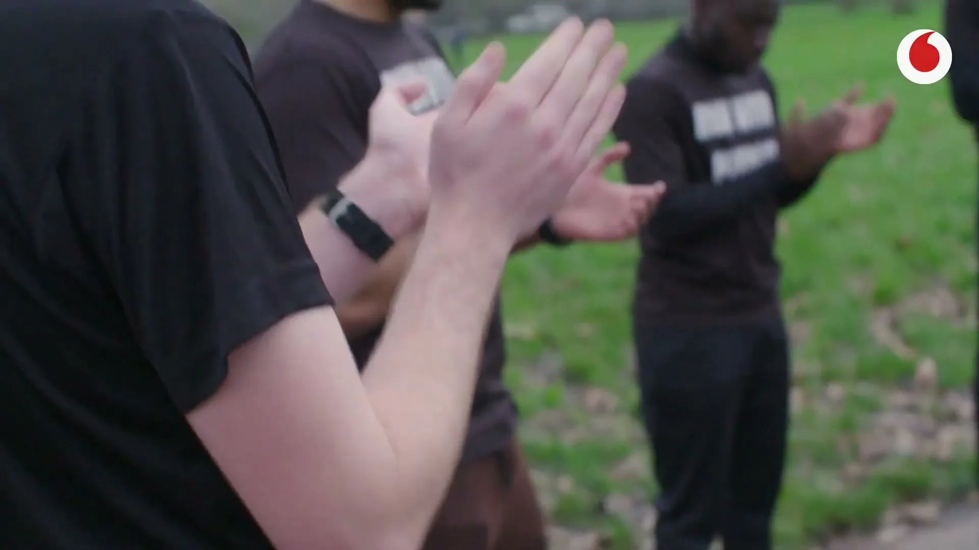 A group of diverse men in black t-shirts and pants clap their hands in a park on a cloudy day.
