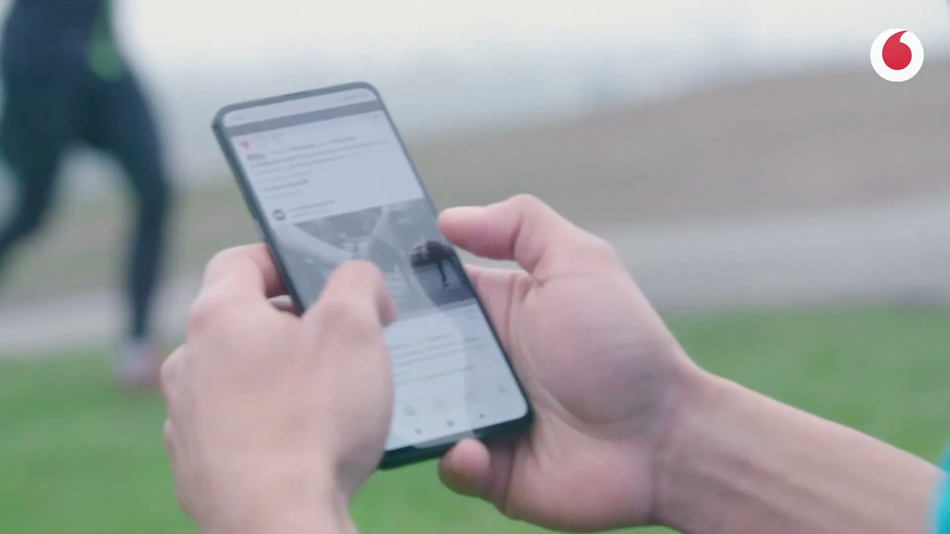 A person's hands hold a smartphone, scrolling through social media, with a blurred background of a person running on grass.