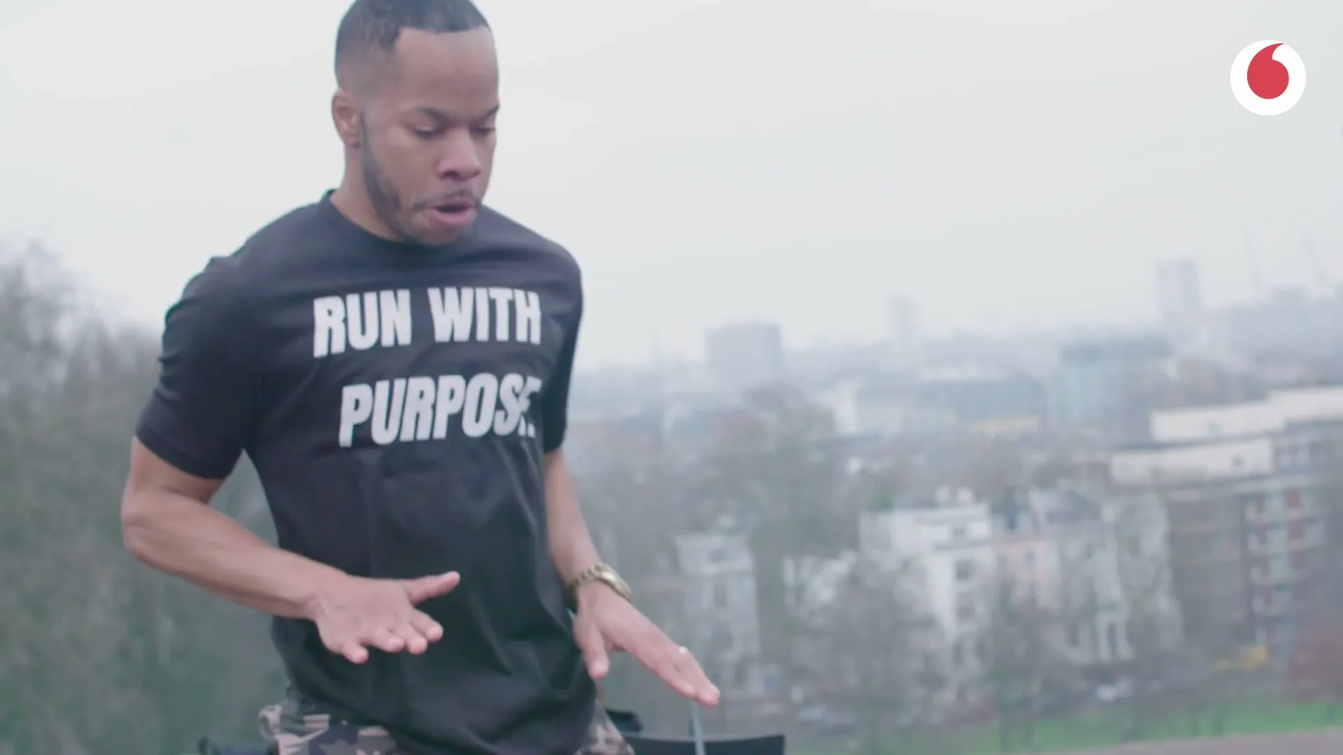 A Black man in his 30s, wearing a black t-shirt that says "RUN WITH PURPOSE", is seen from the chest up, looking down and moving his hands as if playing an instrument, with a city skyline in the background on a cloudy day.