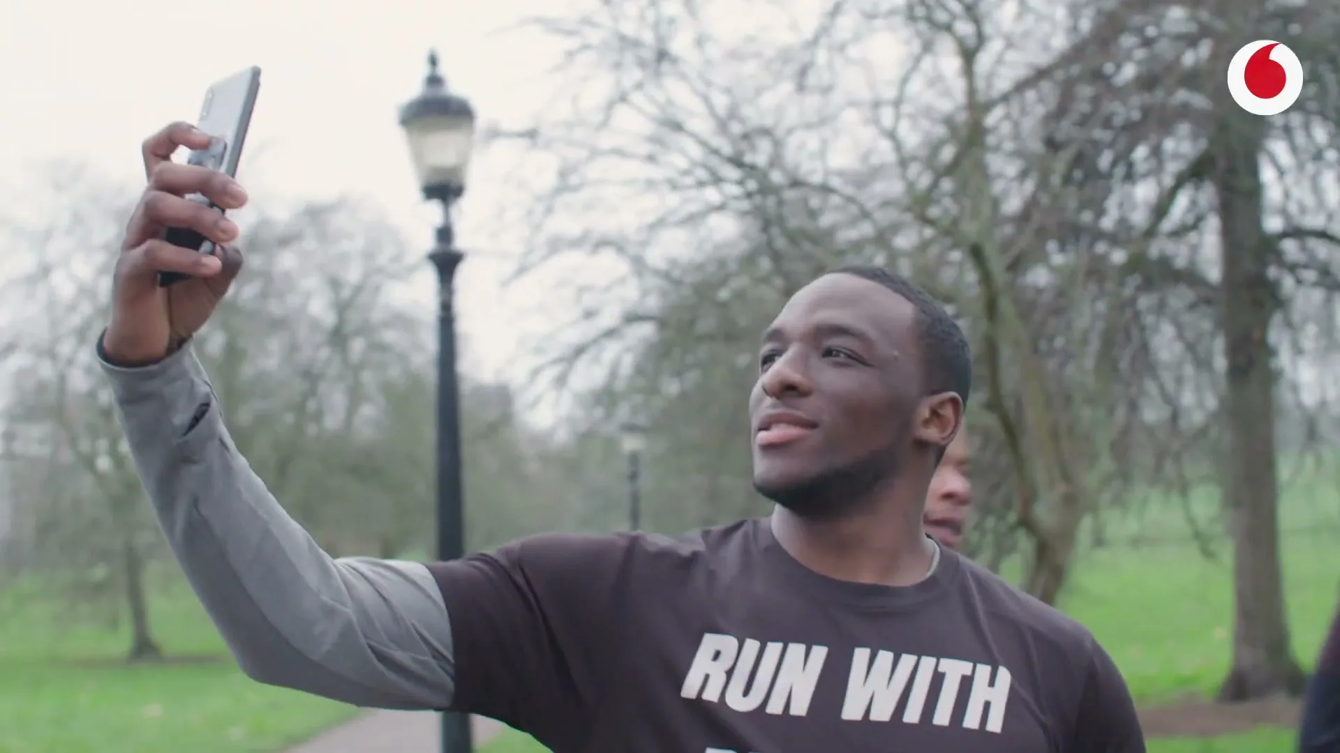 Two Black men in athletic wear take a selfie with a smartphone in a park on an overcast day.