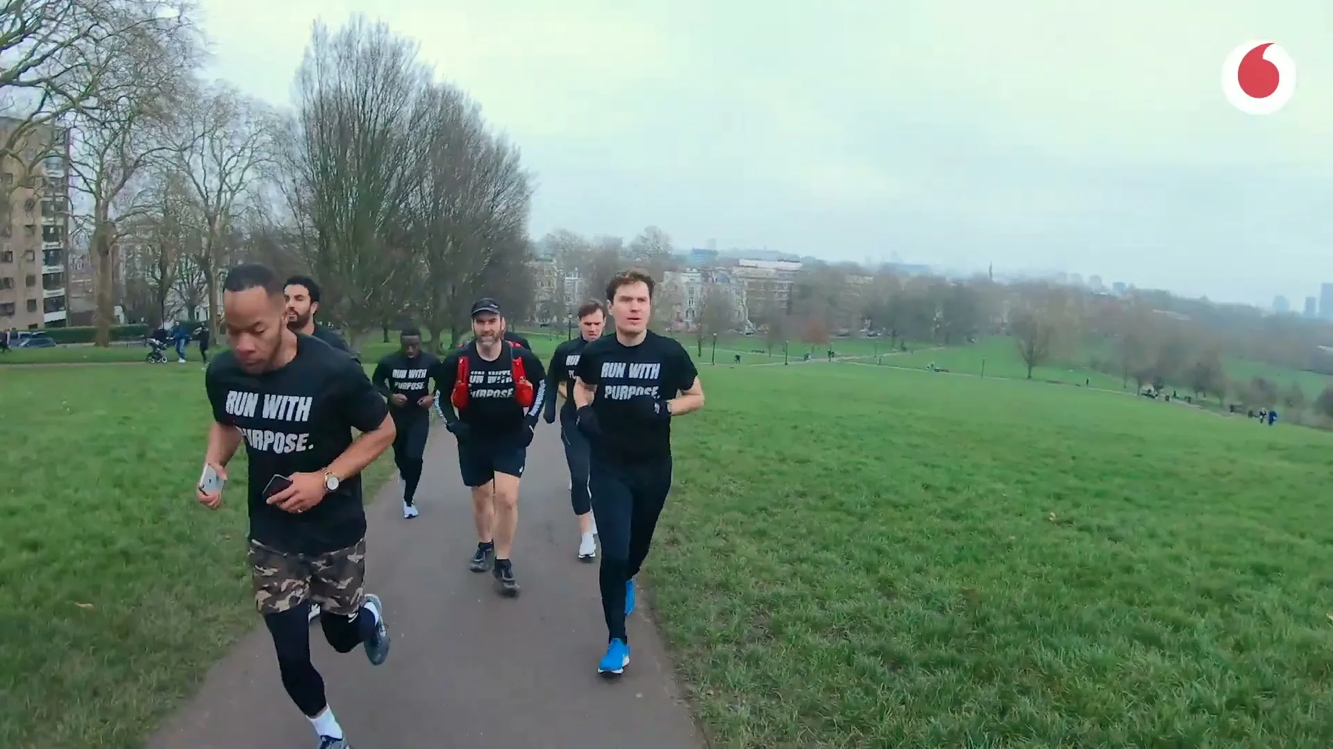 A diverse group of six men in black t-shirts with "RUN WITH PURPOSE" printed on them, run along a paved path in a park on an overcast day.