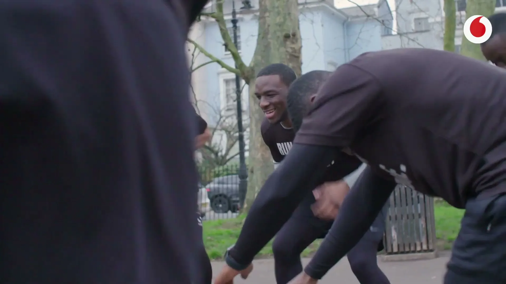 A group of young men in black athletic wear are stretching and laughing together in a park on a cloudy day.