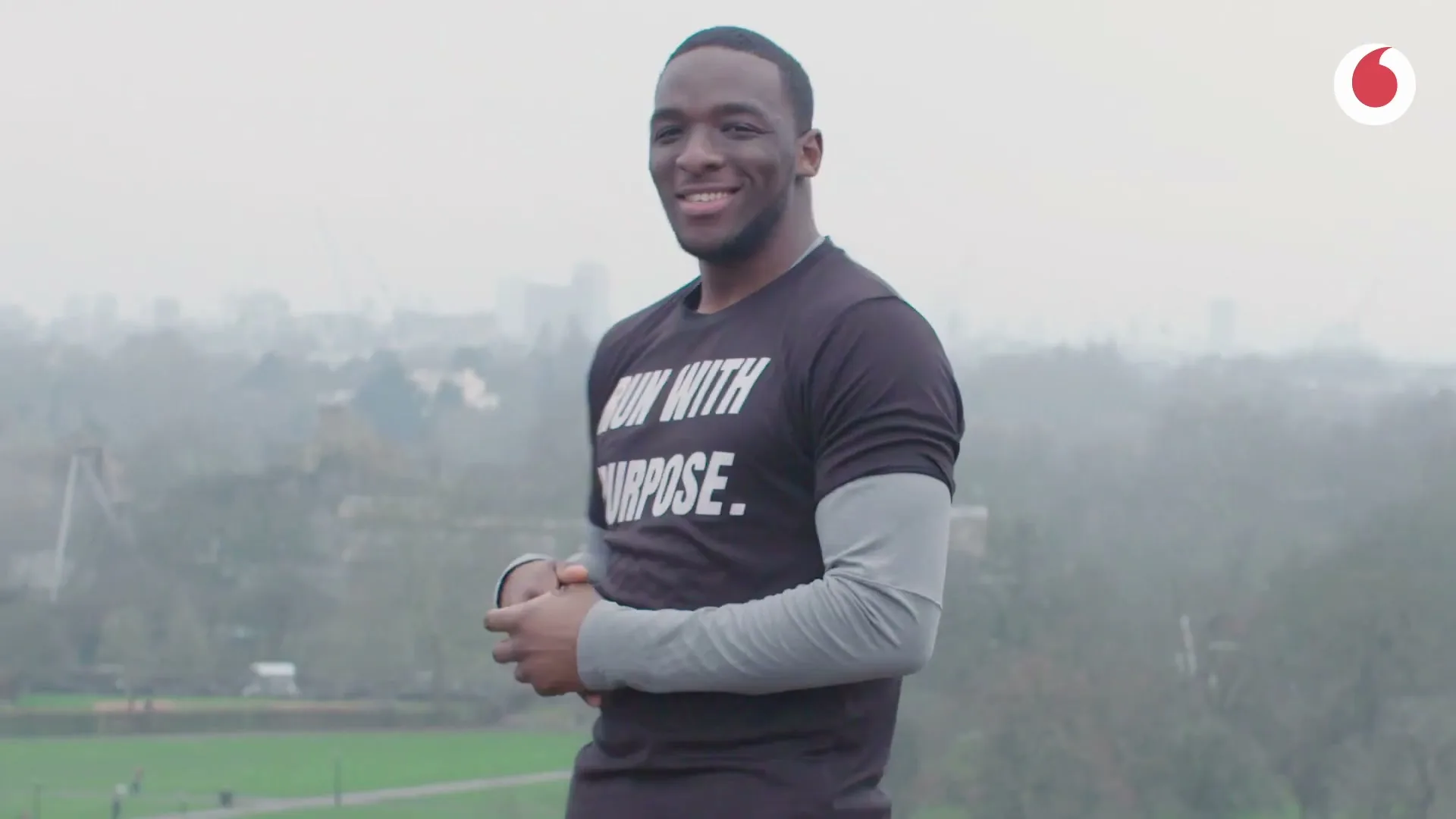 A Black man in his 20s, wearing a black t-shirt that says "RUN WITH PURPOSE", smiles at the camera outdoors on a hill overlooking a city.