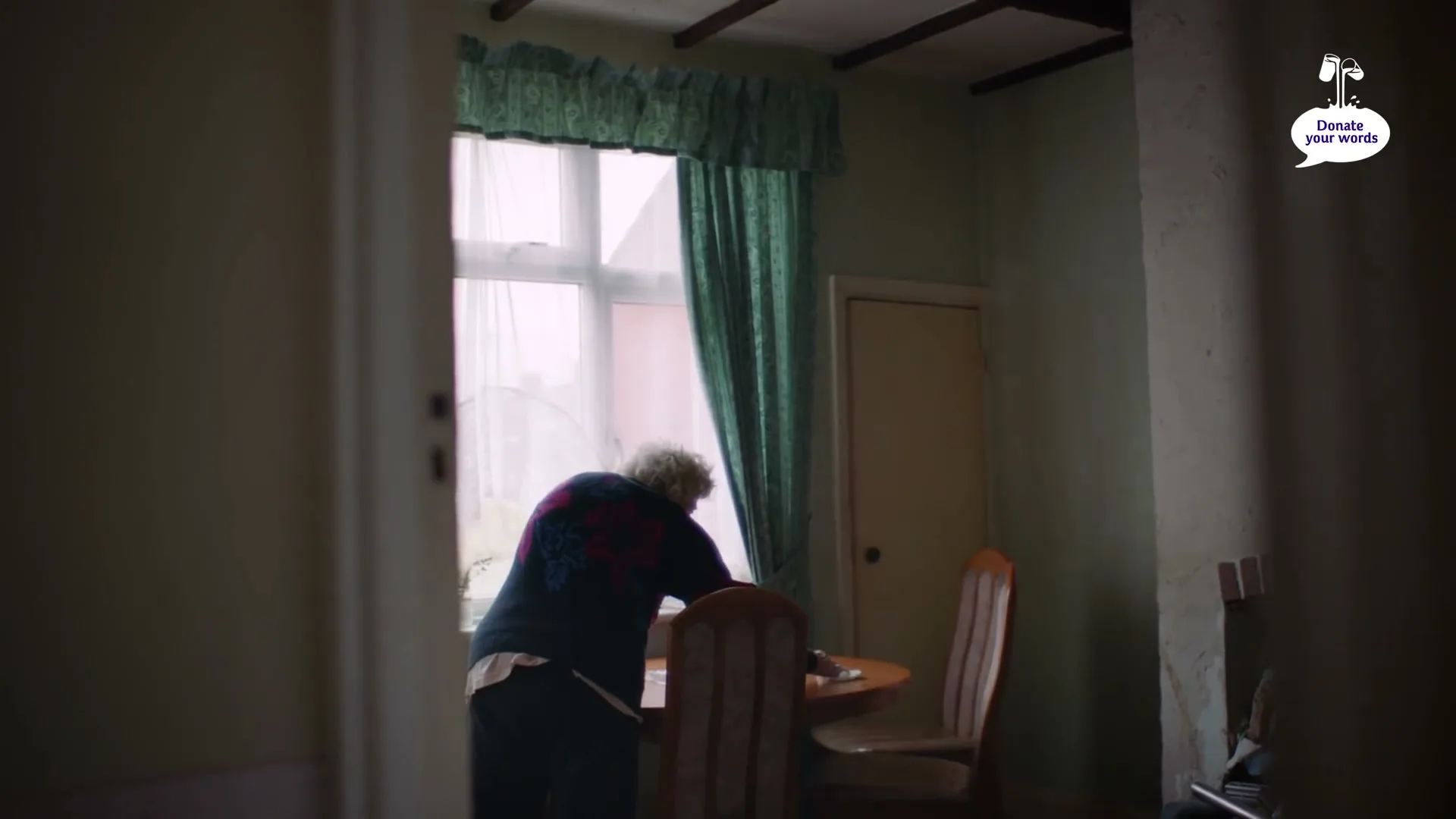 An elderly woman with white hair, wearing a dark blue sweater with red flowers, stands by a window in a dimly lit room, looking out.