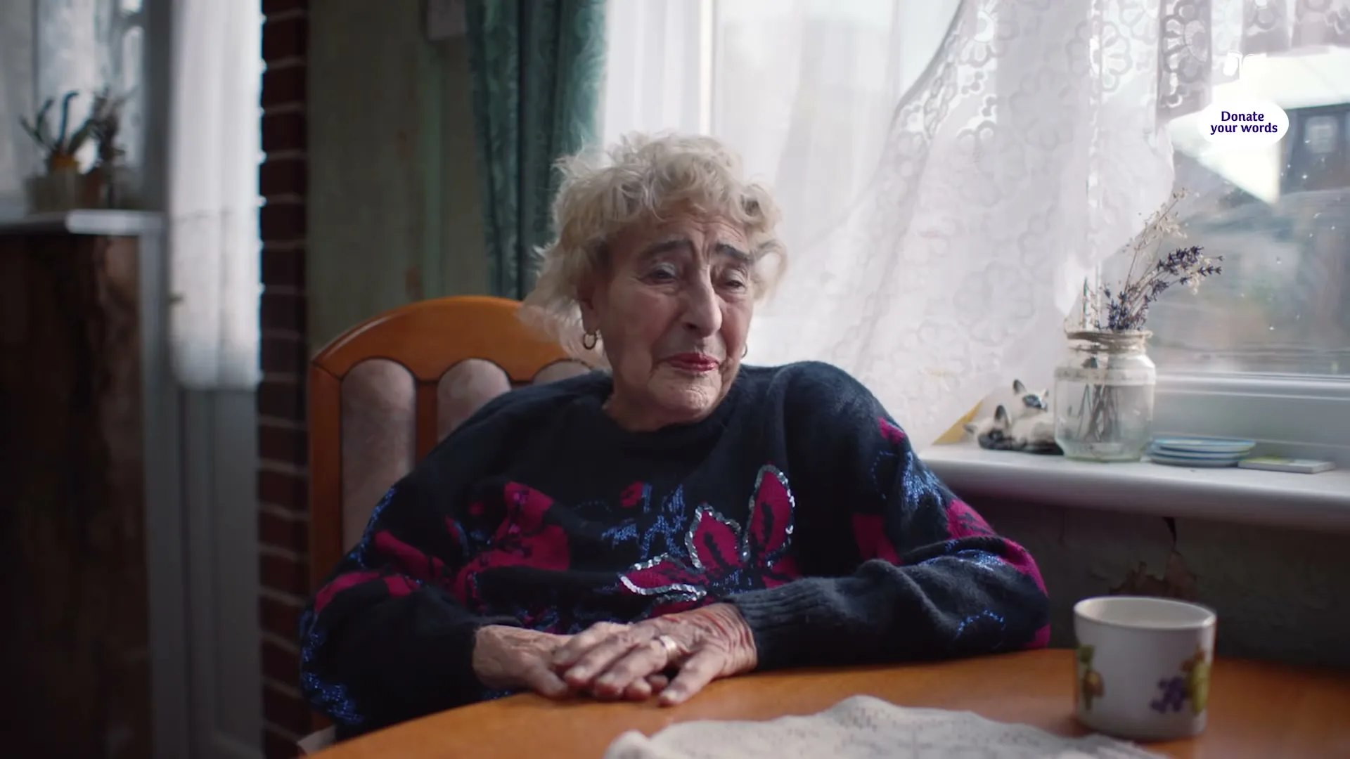 An elderly woman with curly white hair sits at a wooden table, wearing a dark patterned sweater, looking sad in a cozy kitchen.