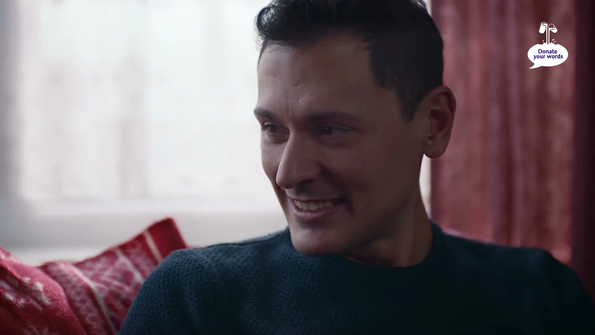 A man in his 30s, wearing a dark blue sweater, smiles and talks while sitting on a couch with a red patterned pillow in a cozy living room.