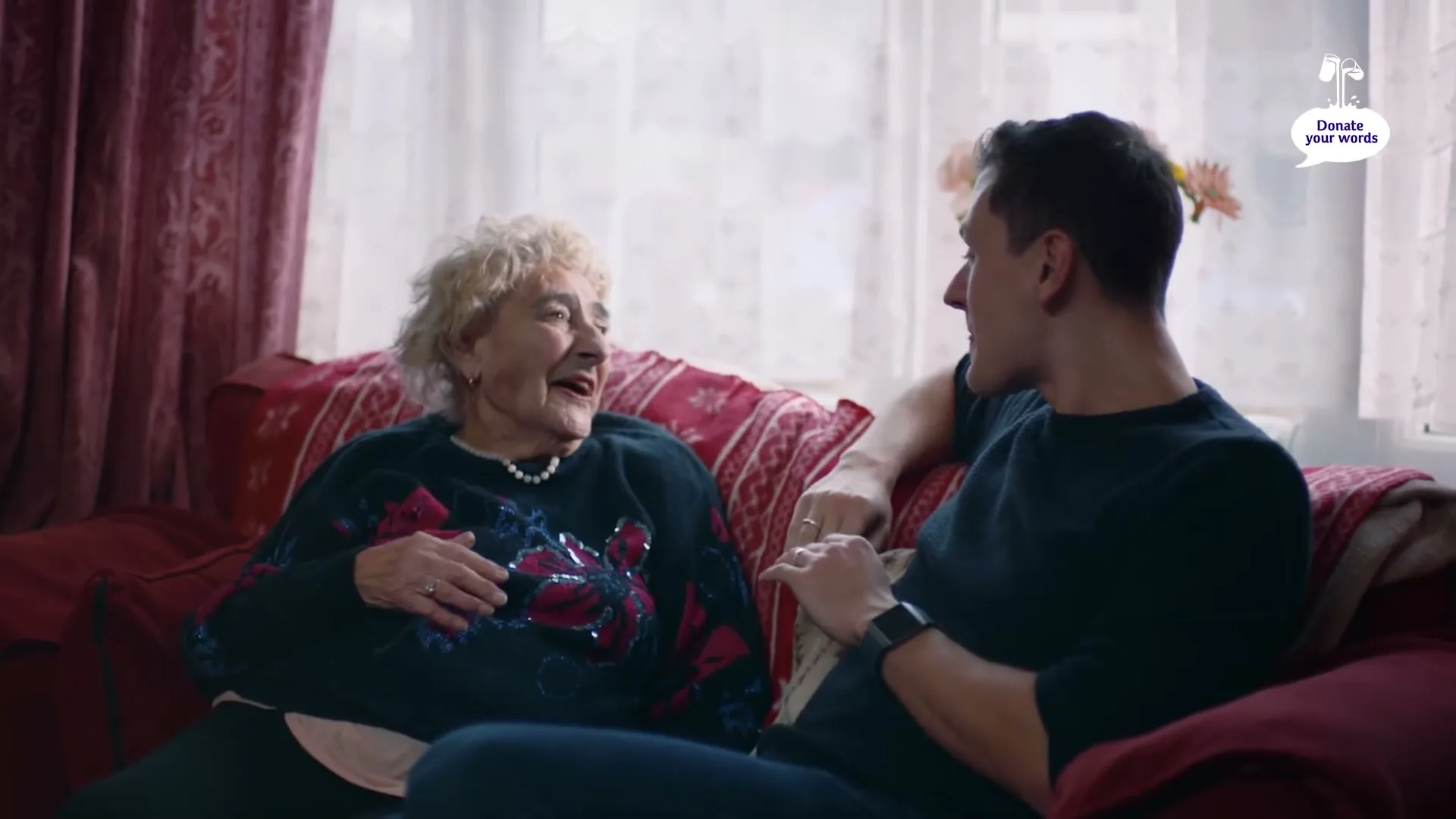 An elderly woman with curly blonde hair and a young man with dark hair sit on a red couch, laughing and talking in a cozy living room.
