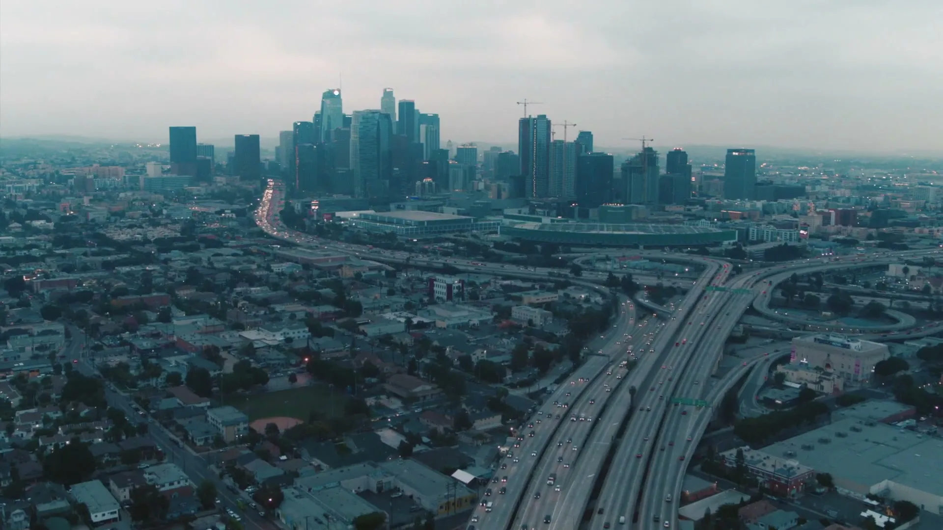 An aerial view of a sprawling city with a complex highway interchange in the foreground and a skyline of skyscrapers in the background under a cloudy sky.