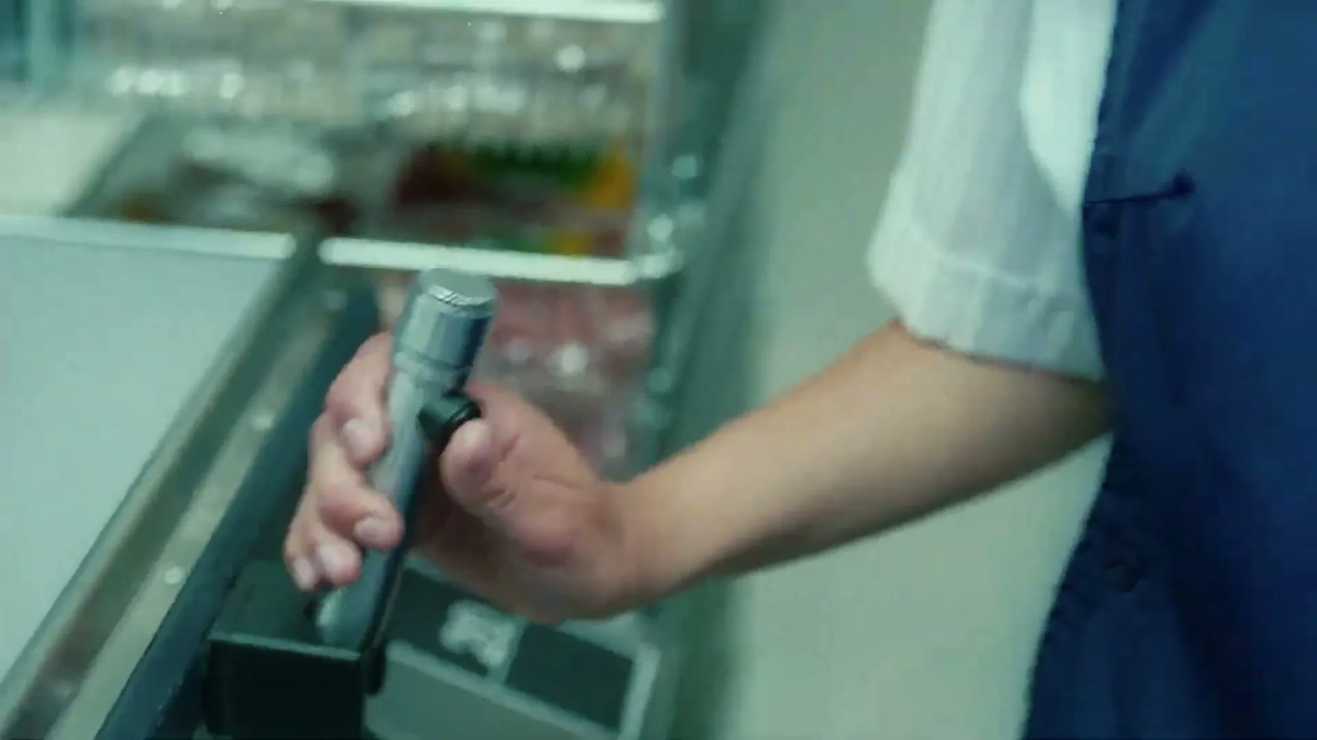 A person in a white shirt and blue apron holds a silver microphone, about to speak into it, in a brightly lit interior.