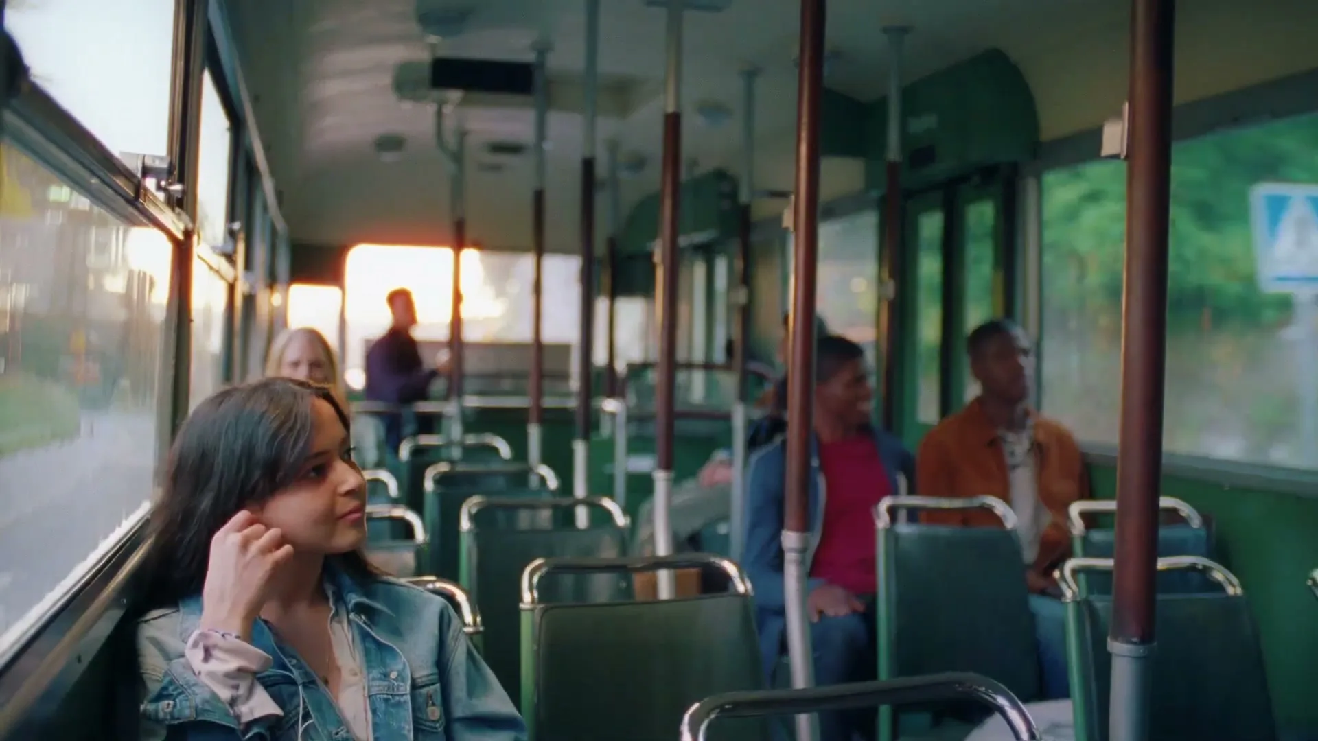 A young woman with dark hair and a denim jacket sits on a bus, looking to her right, as other passengers are visible in the background.