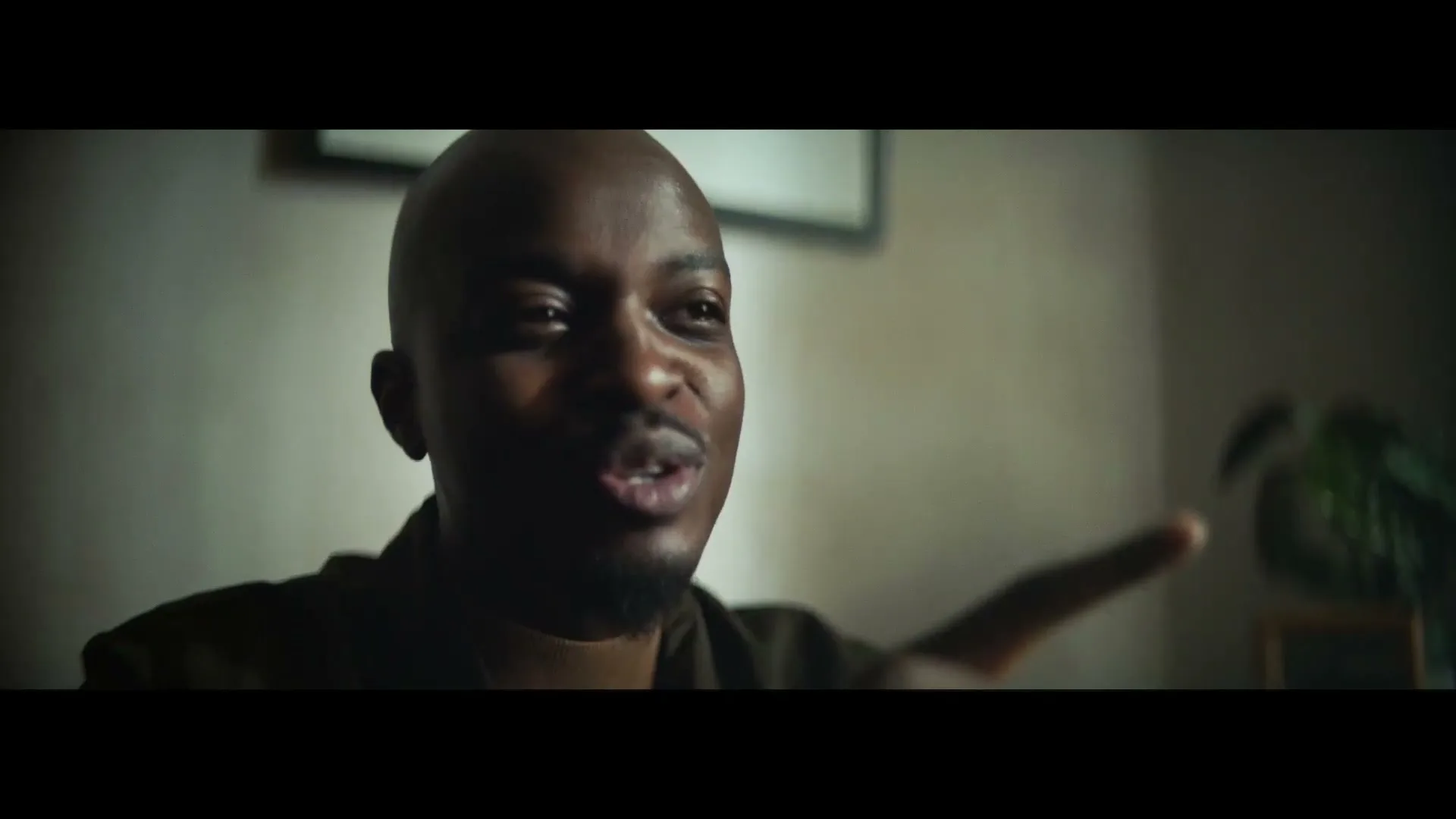 A bald Black man in his 30s, wearing a dark shirt, talks and gestures with his right hand in a dimly lit room.