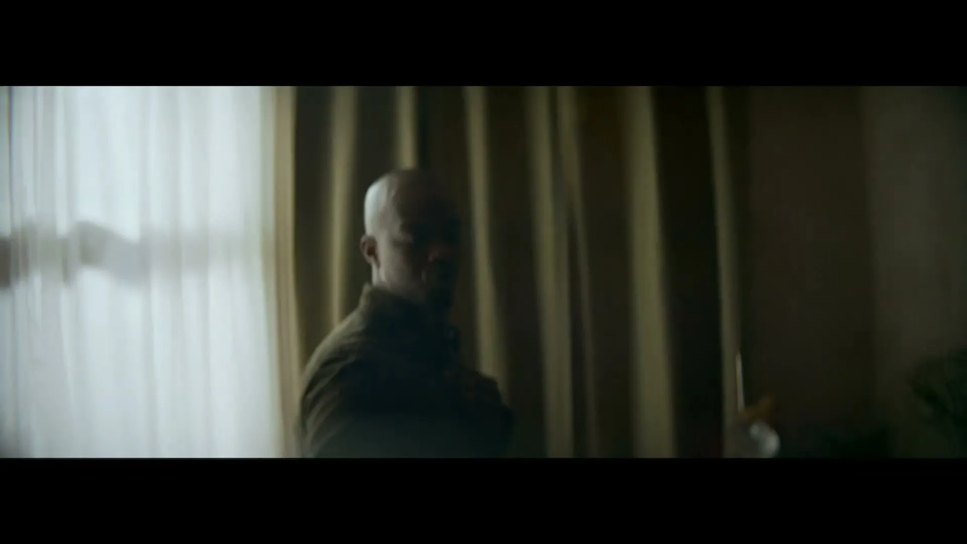 A bald man in his 30s, wearing a dark shirt, turns to face the camera in a dimly lit room with curtains.
