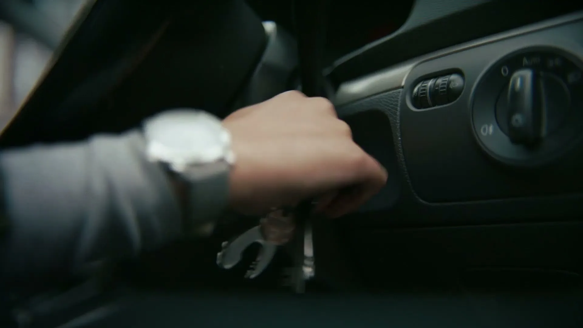 A person's hand with a watch and ring inserts a set of keys into a car's ignition, turning them to start the vehicle.