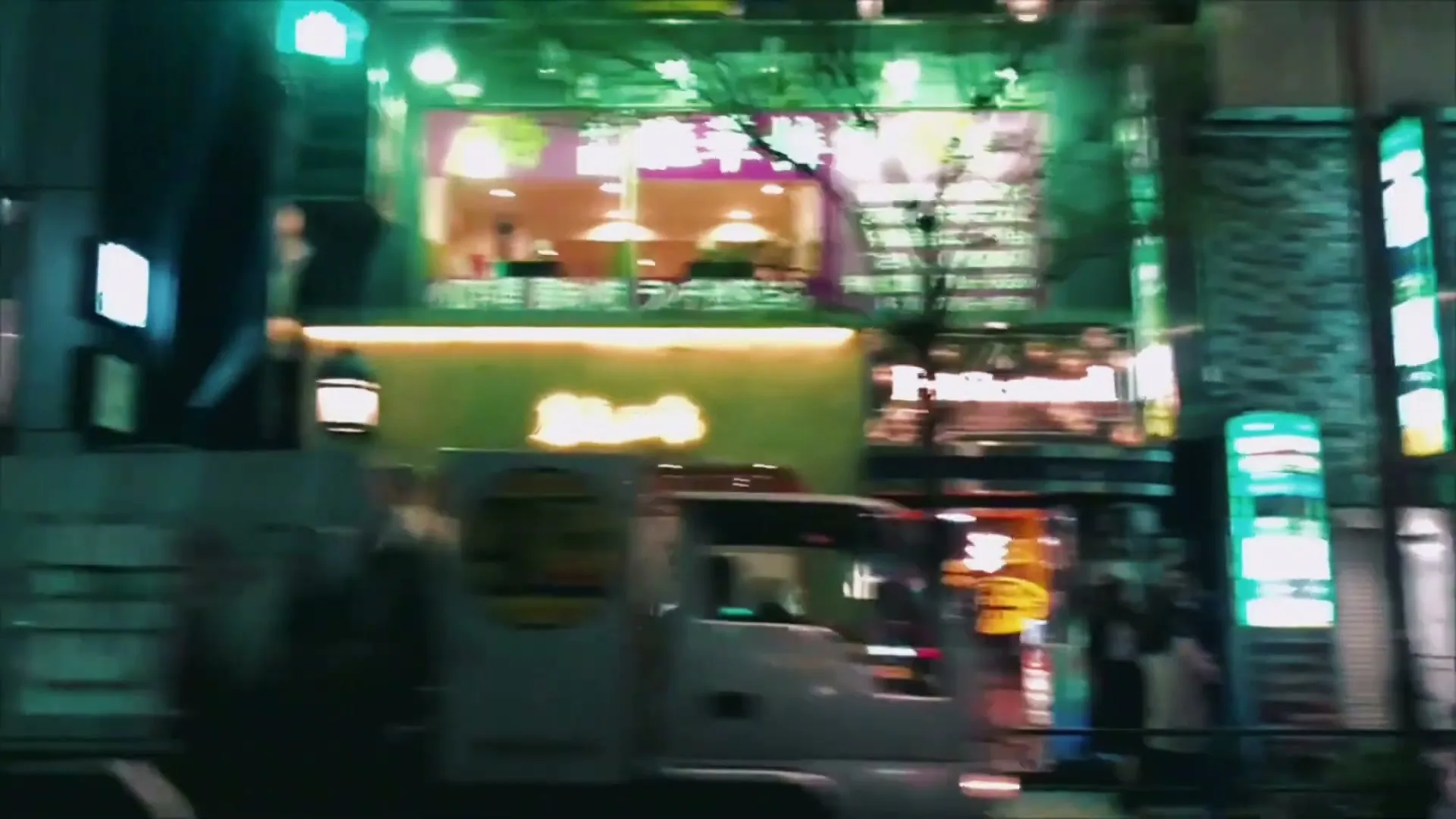 A white truck drives past a brightly lit building with green and purple neon signs in a bustling city at night.