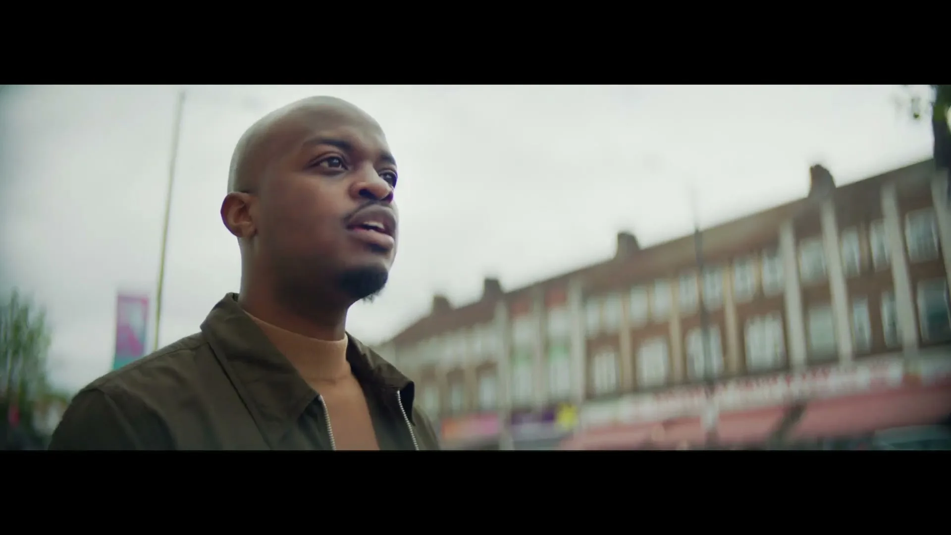 A young man with a buzz cut and beard looks off into the distance on a city street with buildings in the background.