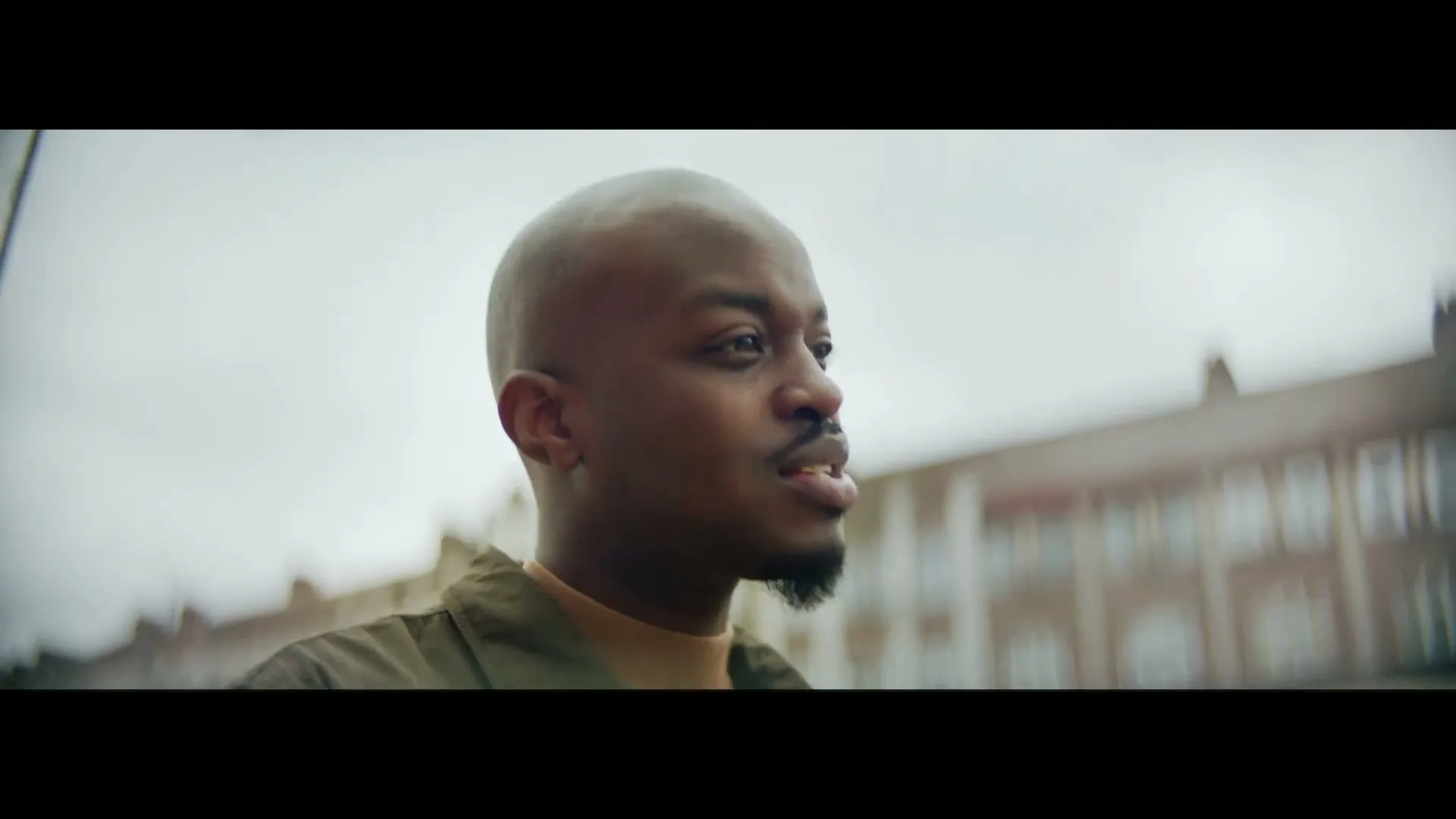A young bald man with a goatee, wearing a green jacket and a tan turtleneck, looks right and speaks on a cloudy day in a city.