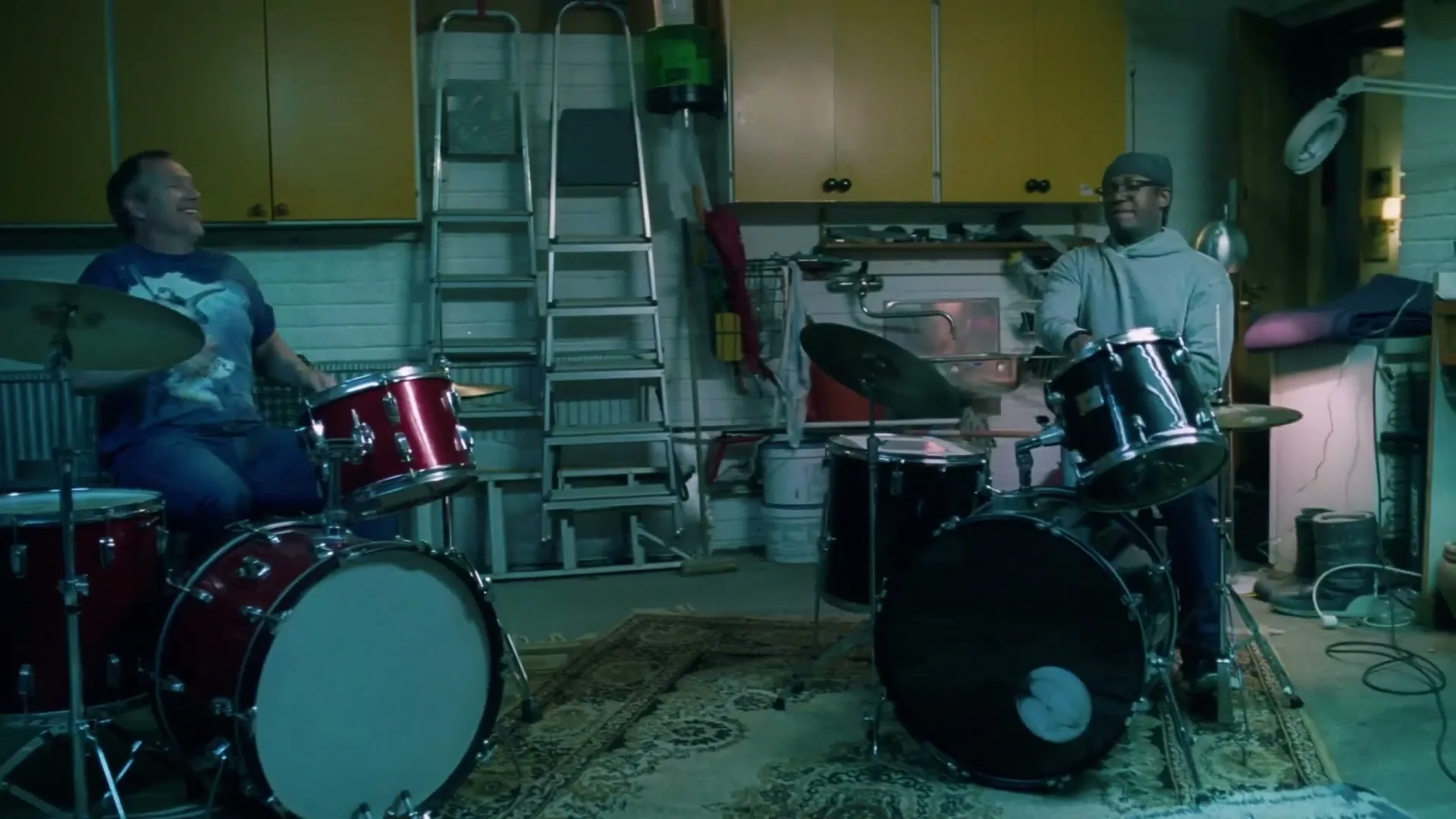 Two men, one white and one Black, play drums in a garage, smiling and laughing, surrounded by tools and storage.