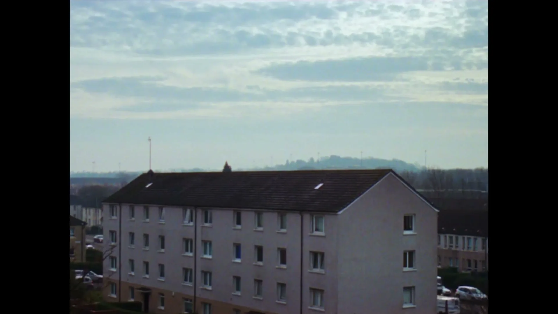 An establishing shot of a residential building with a cloudy sky above, cars parked on the street, and distant hills under a bright sky.
