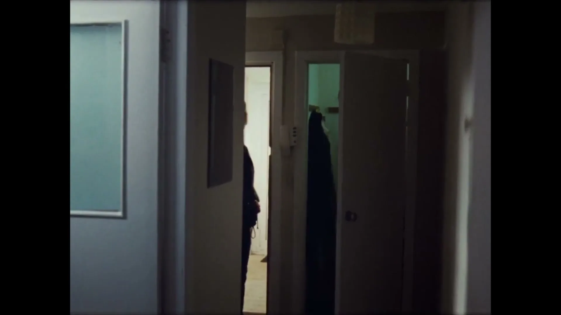 A young woman in a dark jacket and light top walks through a dimly lit hallway, disappearing behind a door.