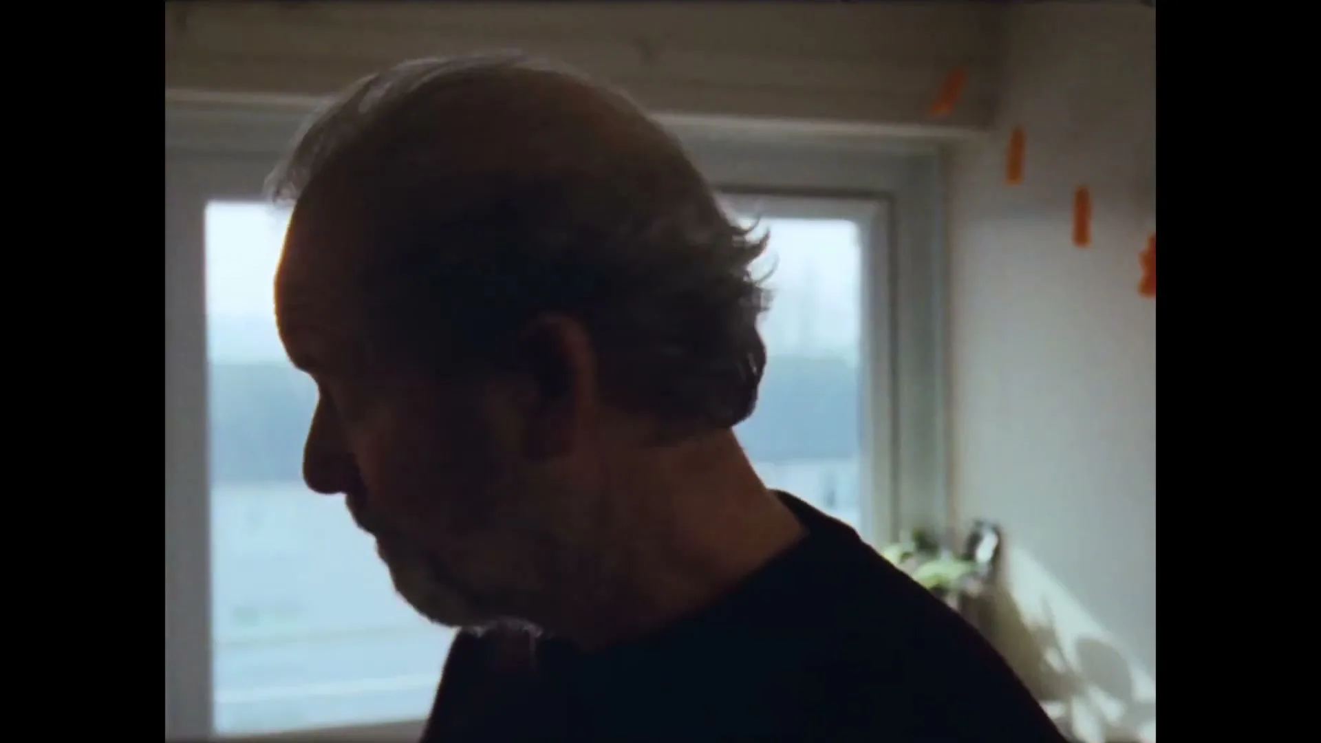 A balding, older man with a beard, wearing a dark shirt, turns his head to the right, looking out a window in a room with orange decorations.