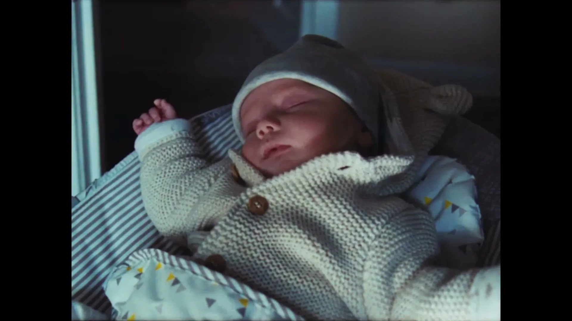 A newborn baby, wearing a grey hat and knitted cardigan, sleeps peacefully in a striped baby bouncer indoors.