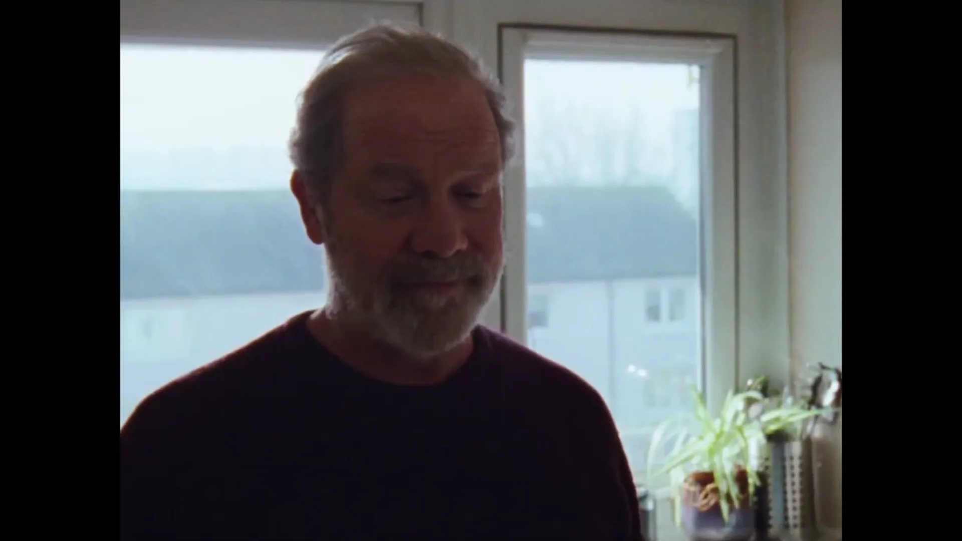 A man with a beard looks down and smiles in a kitchen with a window in the background.