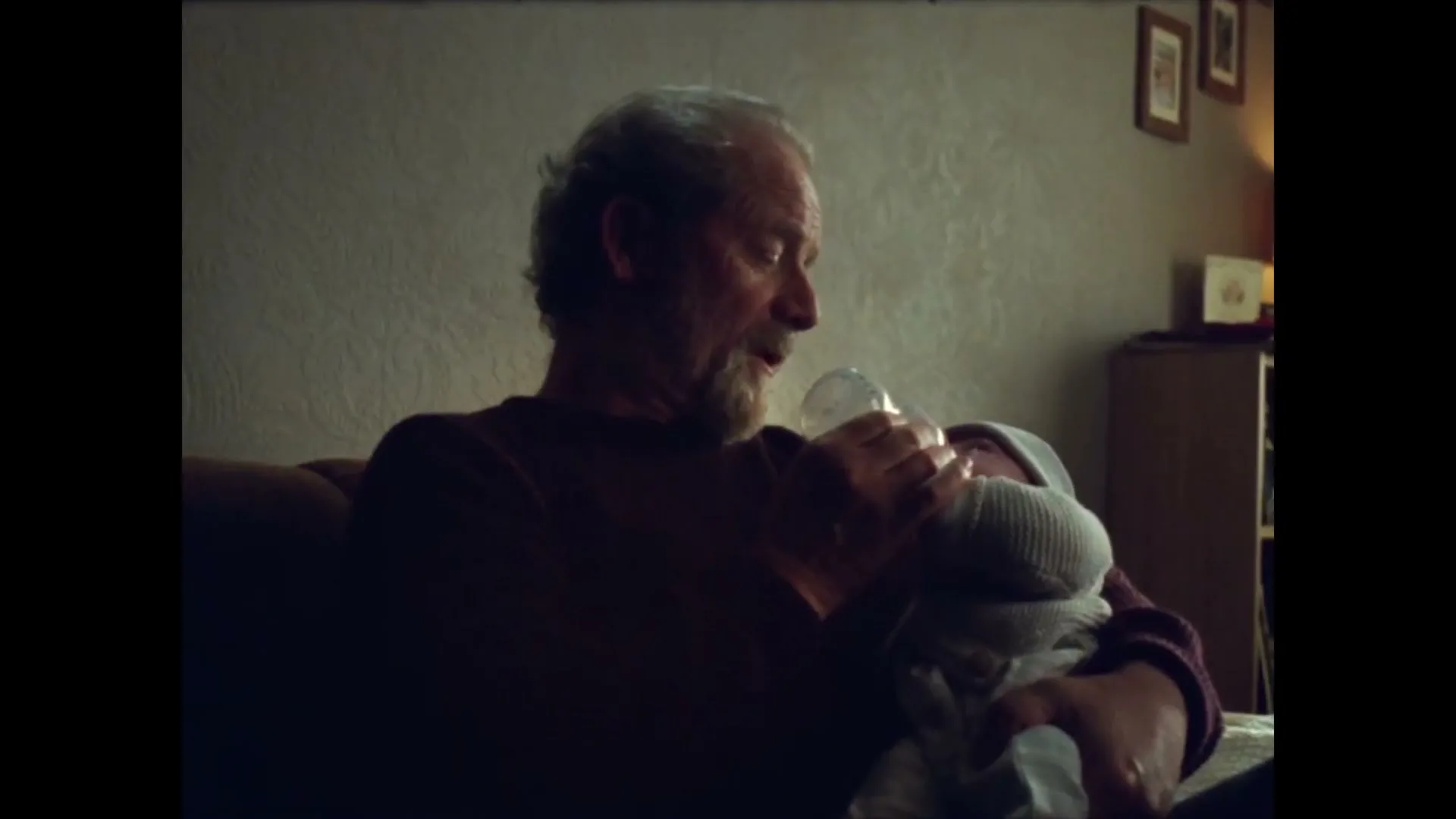 A grey-haired man with a beard sits on a sofa, feeding a baby from a bottle in a dimly lit living room.