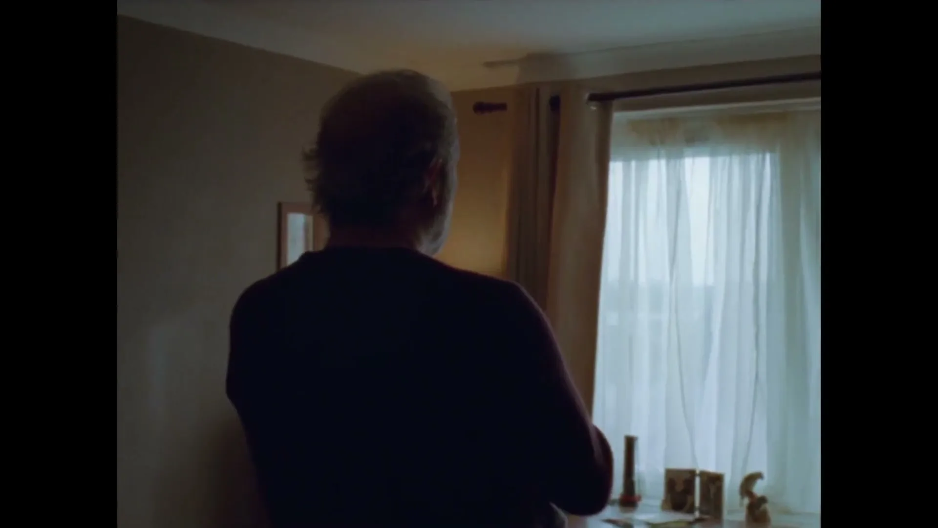 A man with grey hair, seen from behind, holds a baby in a dimly lit room with a bright window in the background.