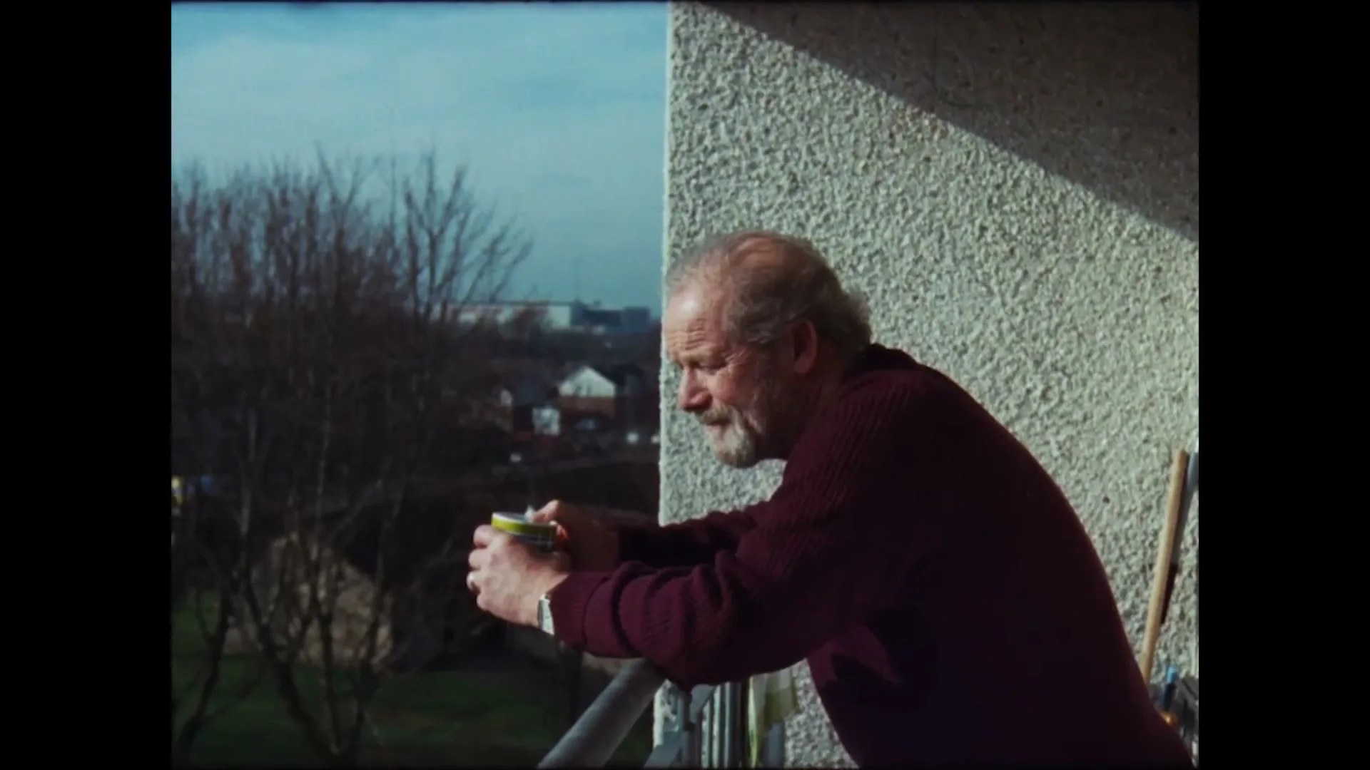 A senior man with a beard, wearing a maroon sweater, leans on a balcony railing, holding a cup and looking out at a suburban landscape.