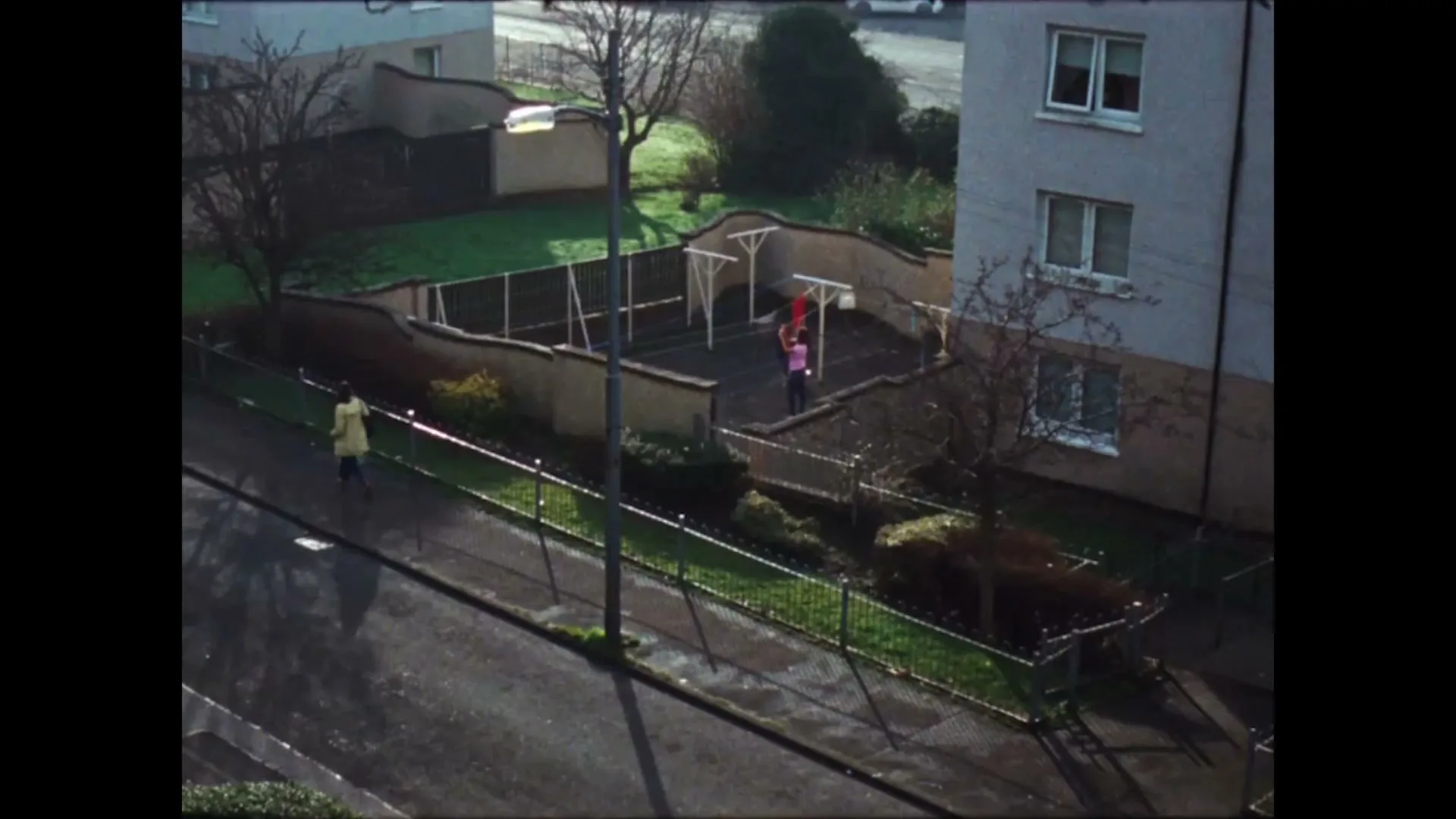 An aerial view shows a child in a yellow jacket walking on a wet street, while two other children play in a fenced-in playground next to an apartment building.