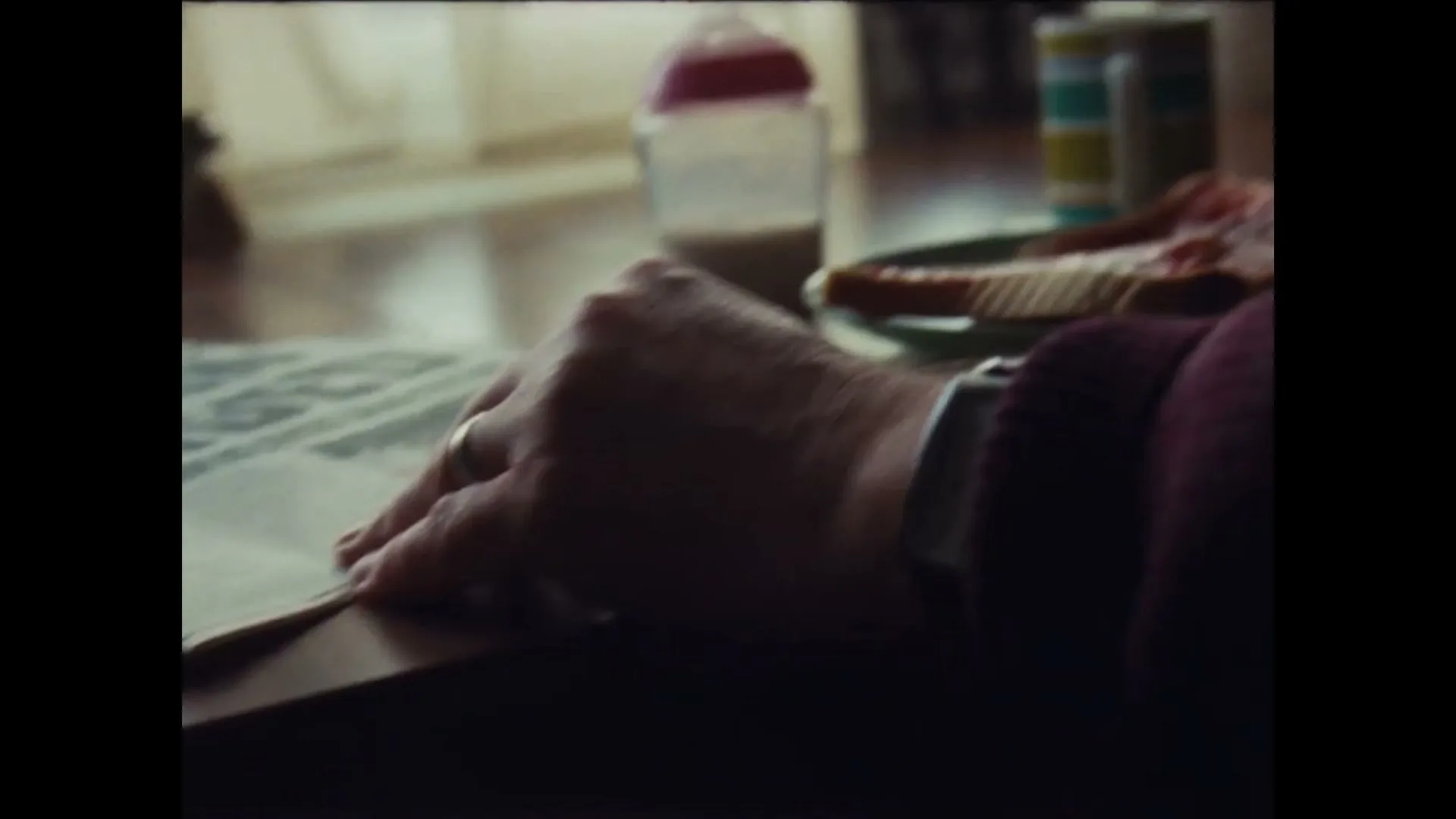 A person's hand with a wedding ring and watch rests on a newspaper on a wooden table, with a baby bottle and toast in the background.