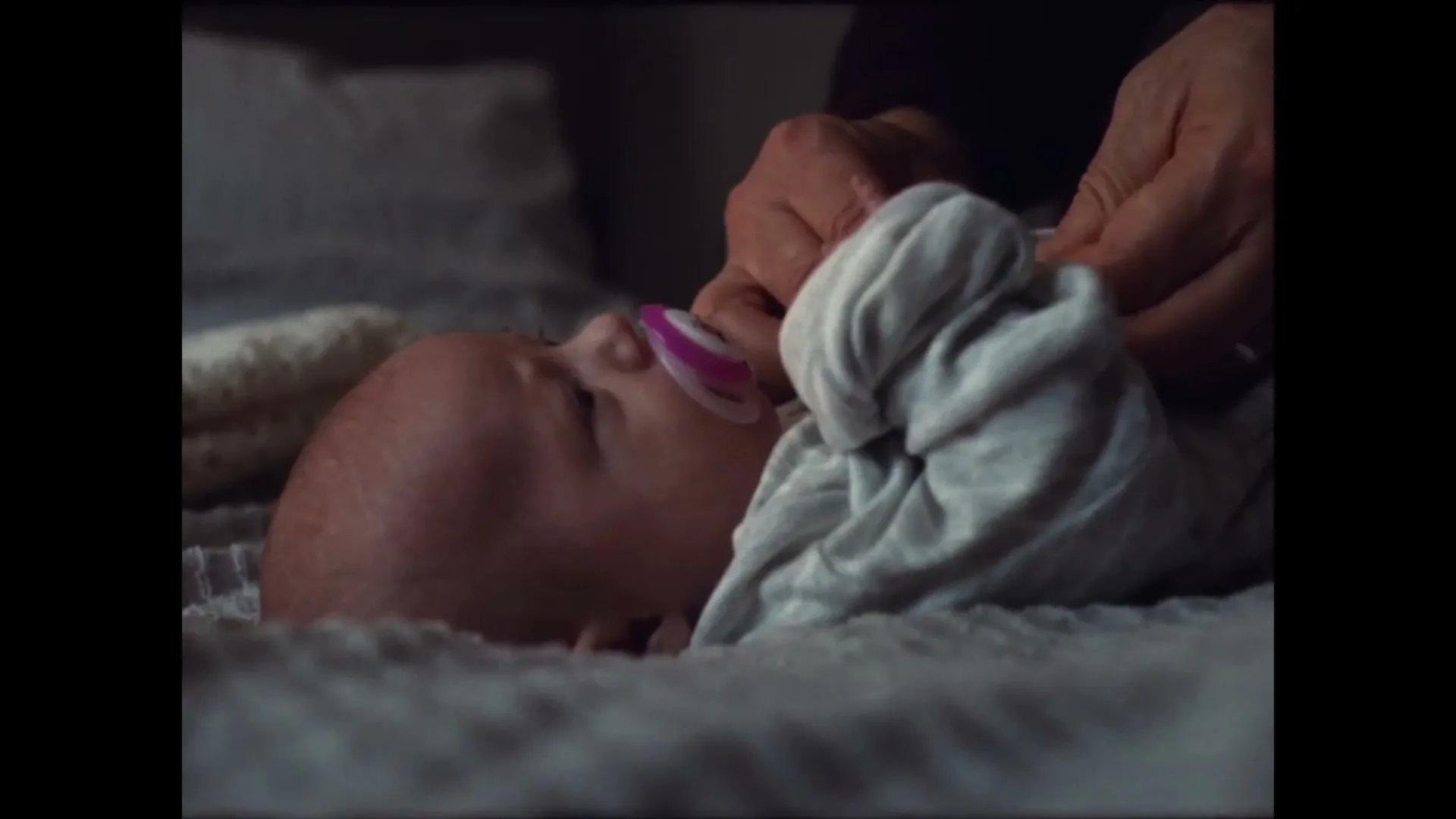 A baby with a pacifier lies on a bed while an adult's hands adjust its clothing in a dimly lit room.