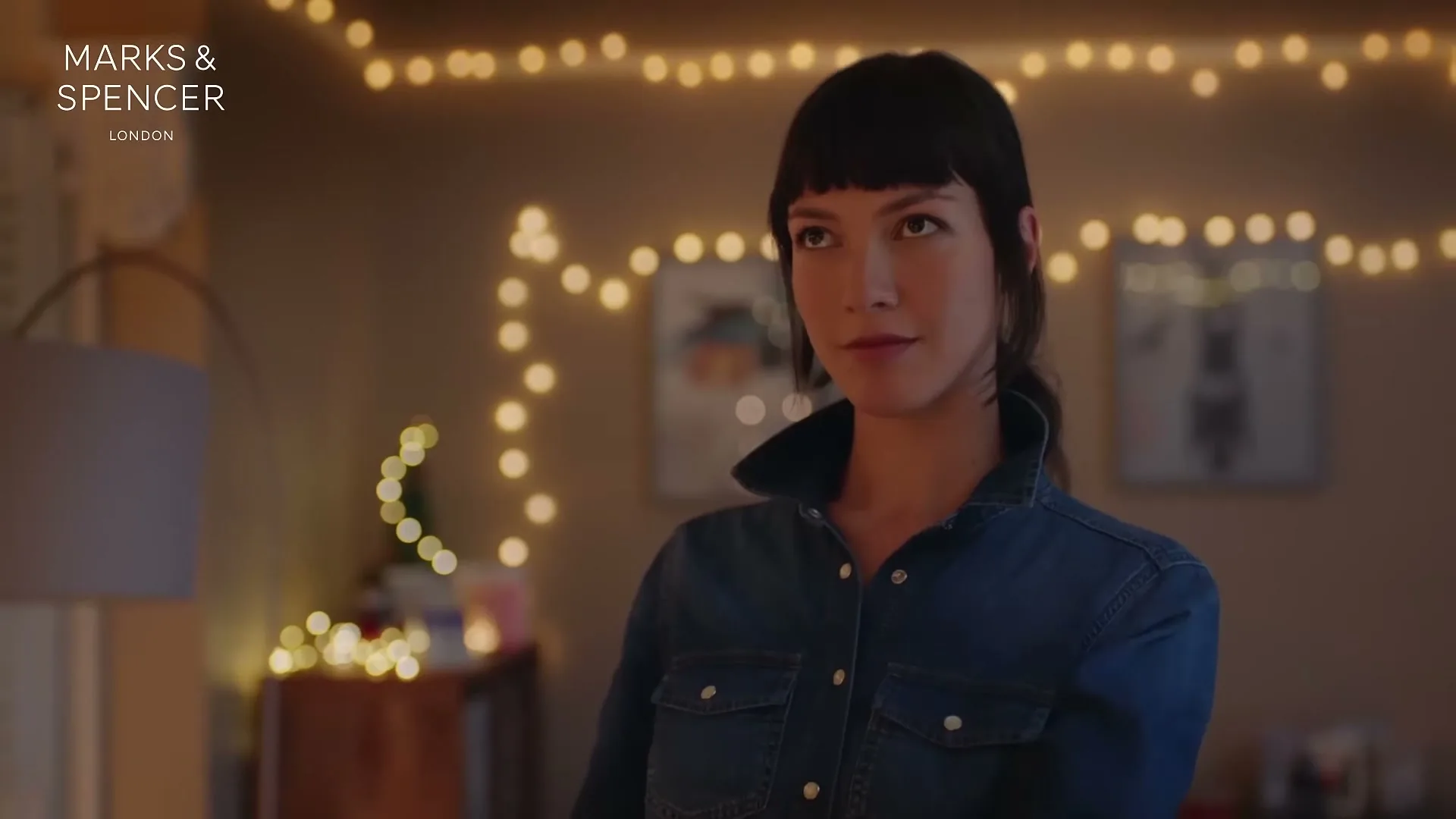 A young woman with dark hair and a denim shirt looks around a cozy room decorated with string lights, as if searching for something.
