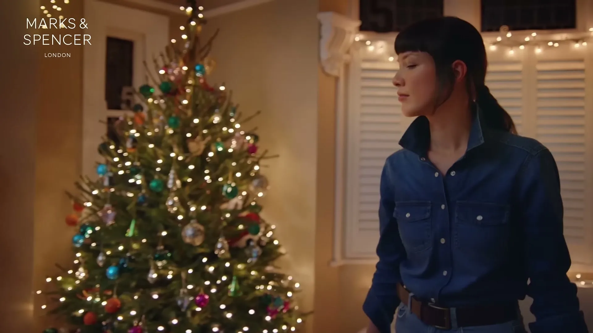 A young woman in a denim shirt looks around a festive living room with a decorated Christmas tree.