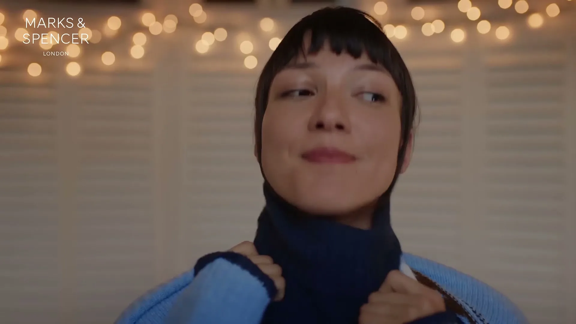 A young woman with dark hair pulls a dark blue turtleneck sweater over her head, then smiles, in a cozy room with string lights.