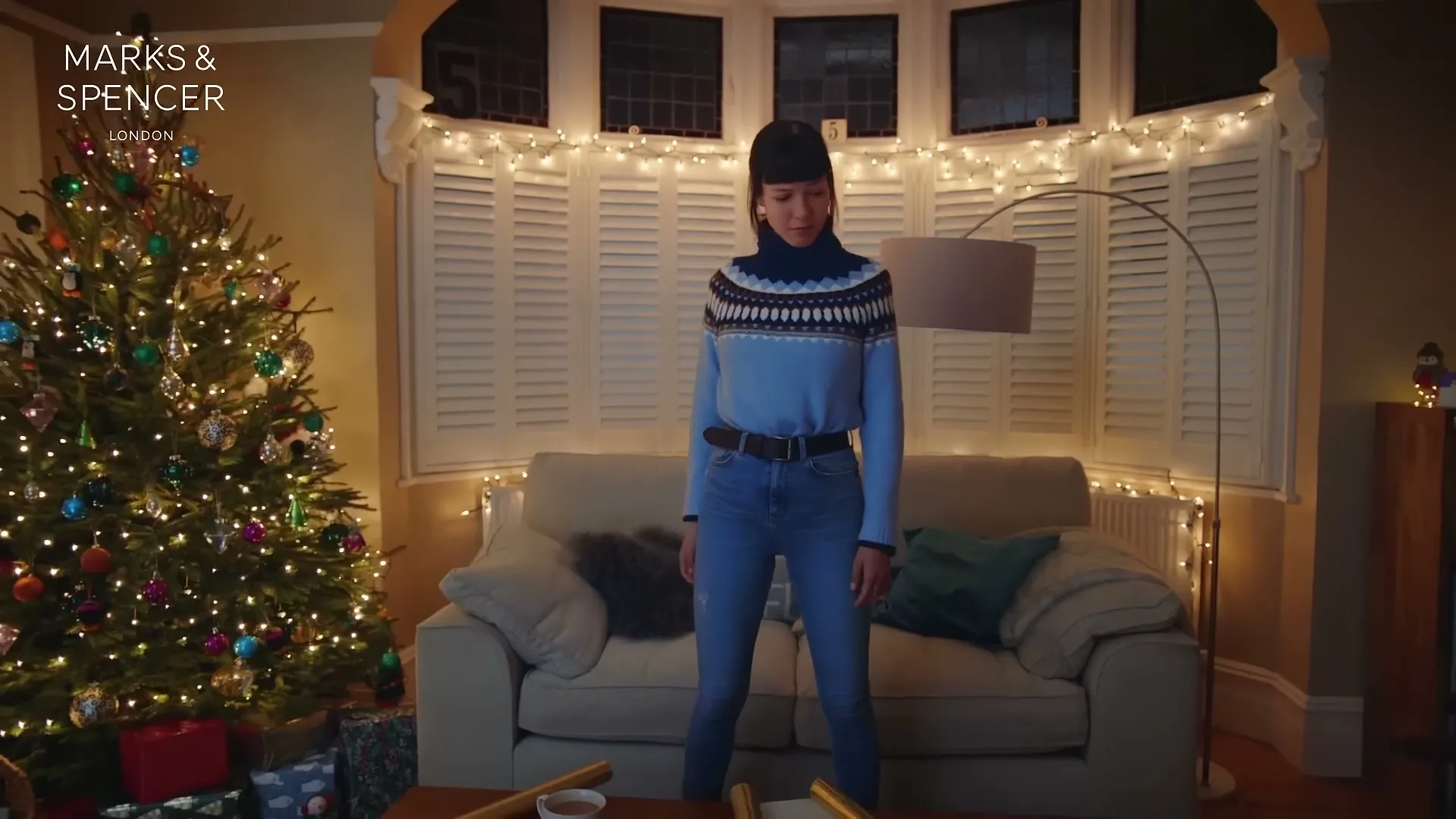 A woman in a blue patterned jumper and jeans dances in a living room decorated for Christmas, with a Christmas tree and fairy lights.