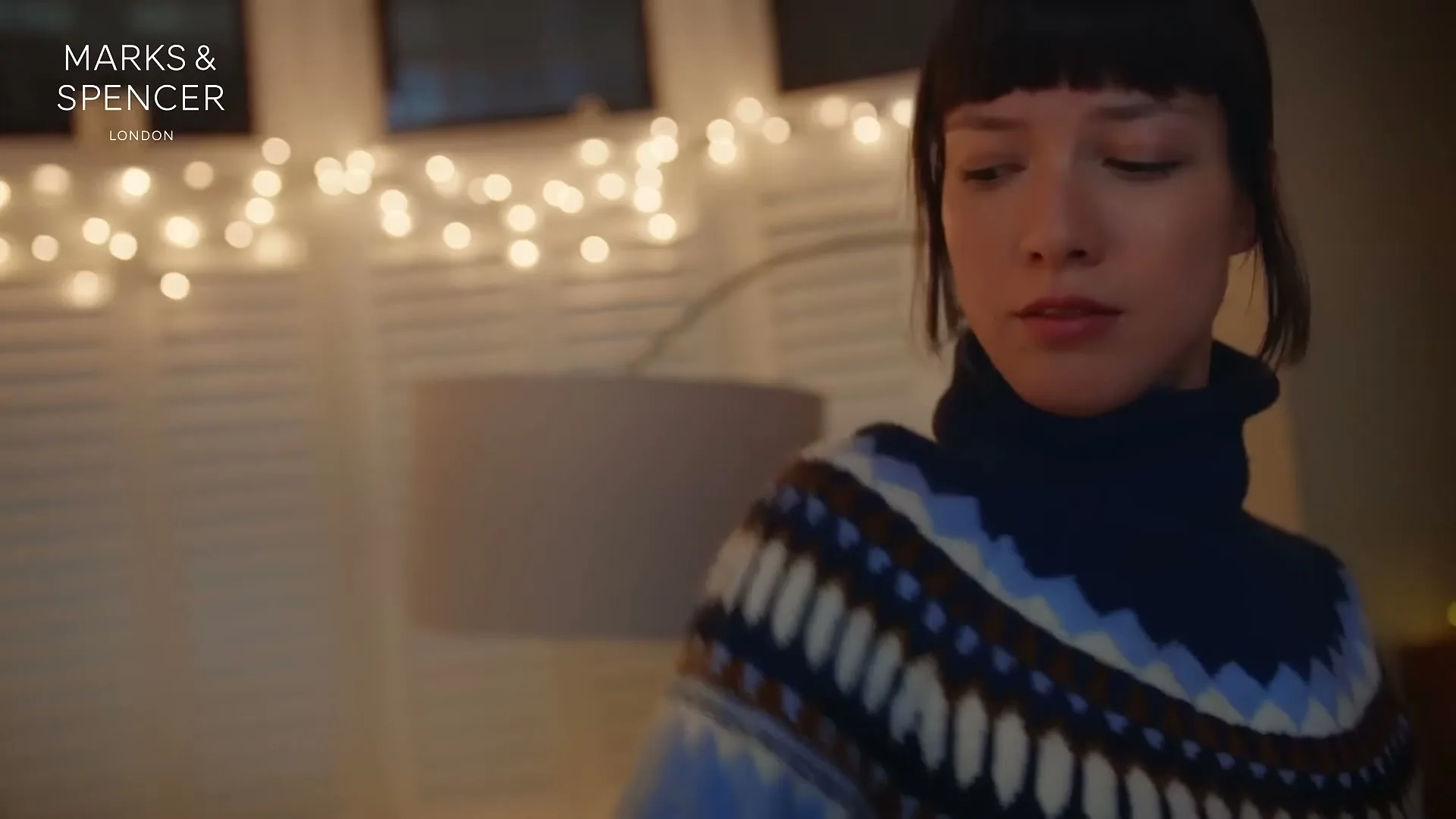 A young woman with short dark hair wears a blue and white patterned turtleneck sweater, looking down in a cozy room with string lights.