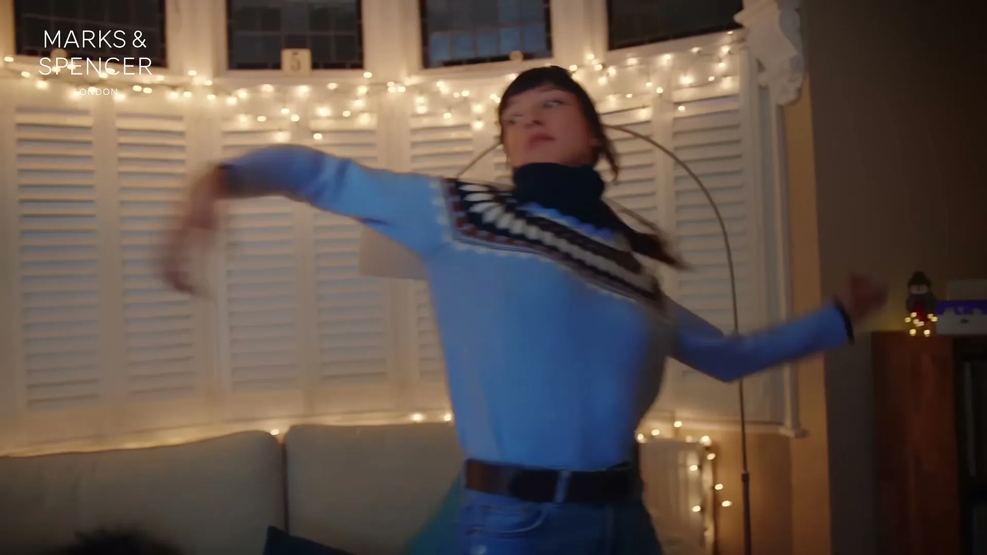 A young woman with long dark hair, wearing a blue patterned jumper, dances energetically in a living room decorated with Christmas lights.