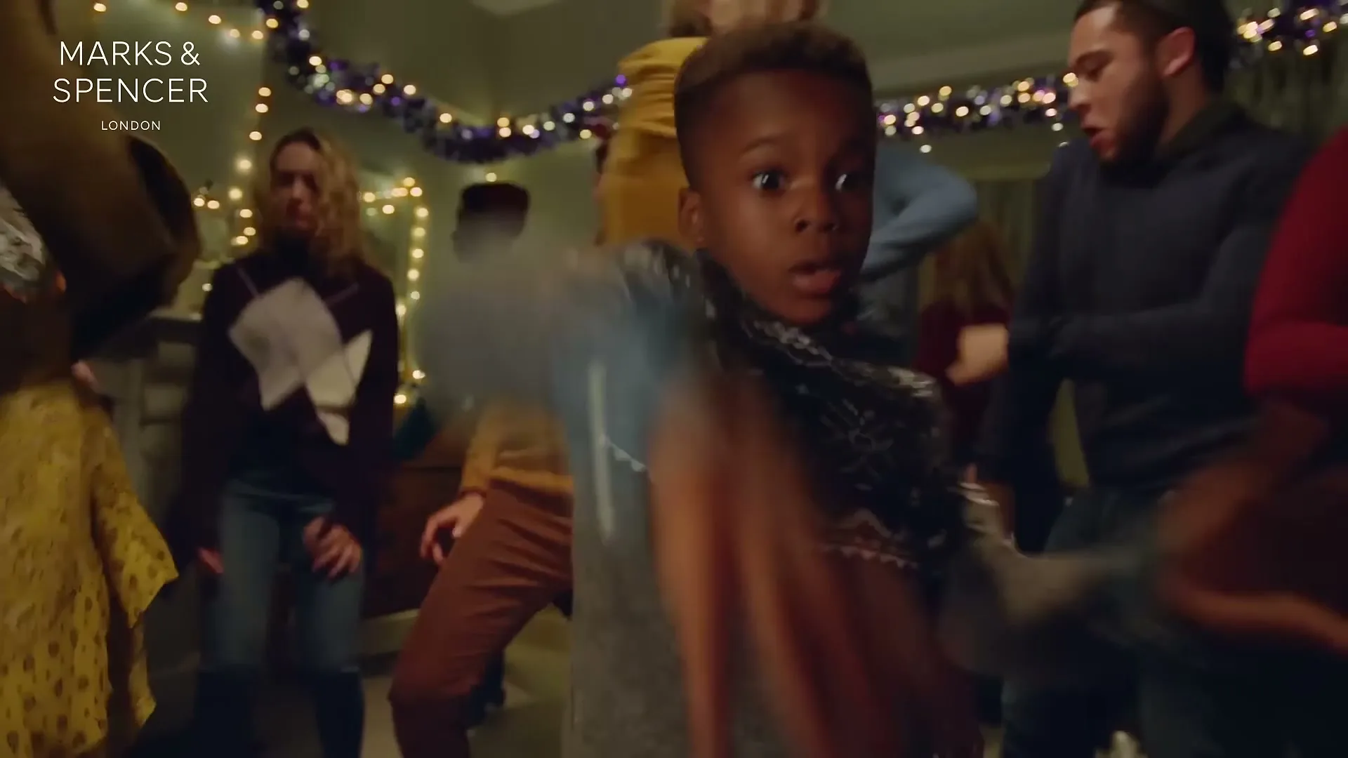 A young Black boy in a grey patterned sweater dances energetically with a surprised expression, surrounded by other people dancing at a festive Christmas party.