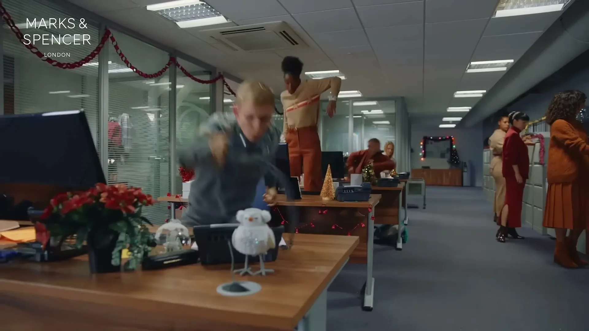 A young man in a Christmas jumper jumps on a desk in an office decorated for Christmas, while colleagues dance in the background.