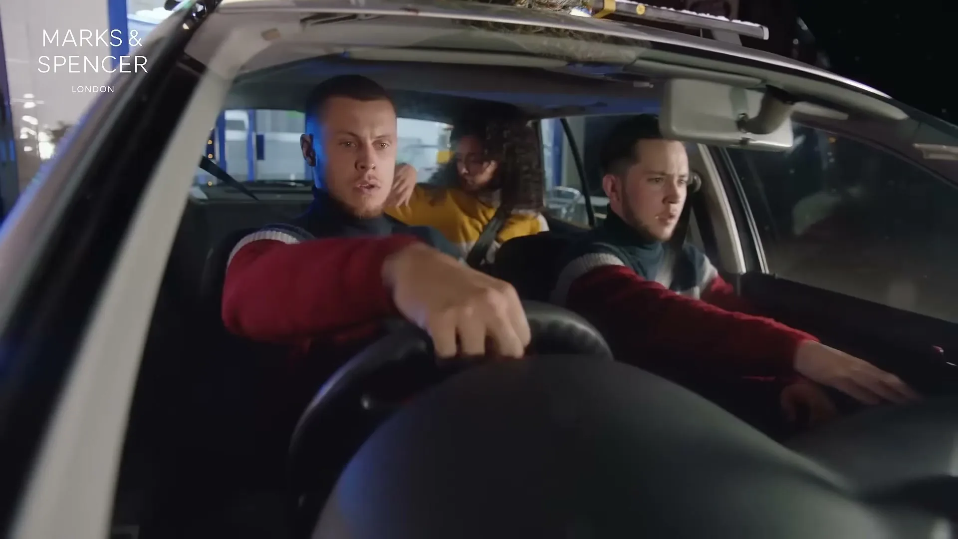 Three young adults, two males in the front and one female in the back, are driving in a car at night, looking surprised.